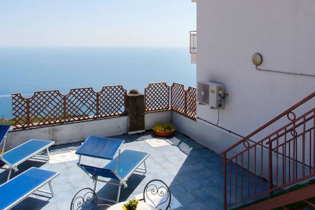 A rooftop terrace with blue sun loungers, a potted plant, and a view of the sea.