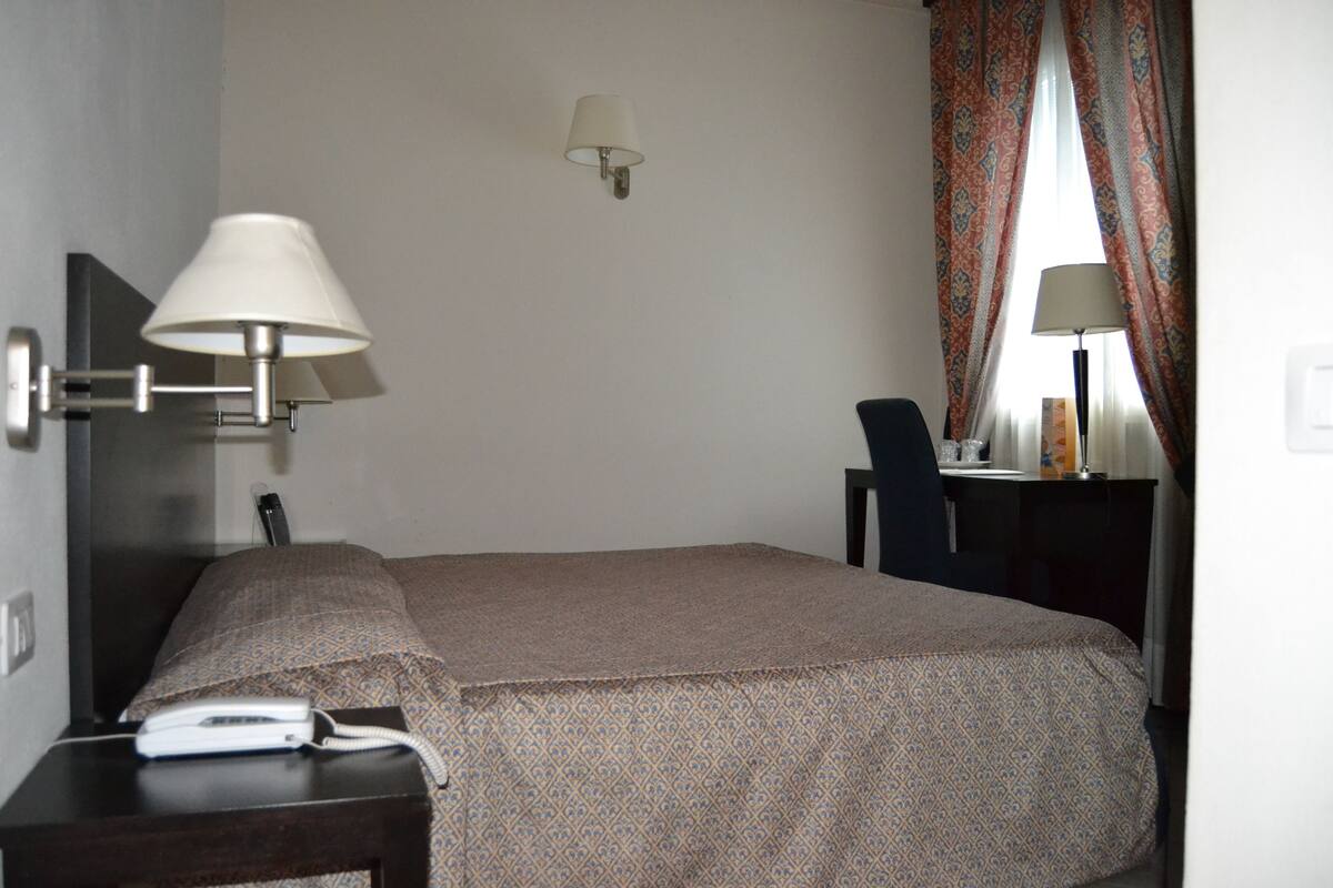 A hotel room with a bed, bedside table, lamp, and a desk with a chair.