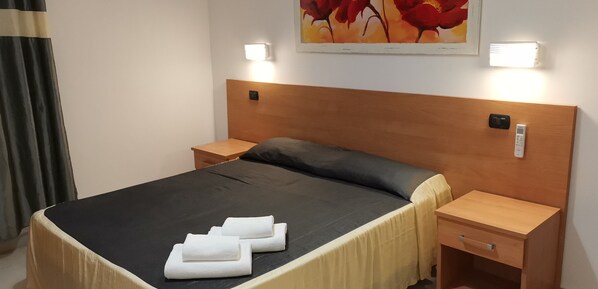 A neatly made bed with a dark bedspread and two folded white towels, wooden nightstands with wall-mounted lamps, and a small wooden desk with a drawer. - Angelica (Rome)