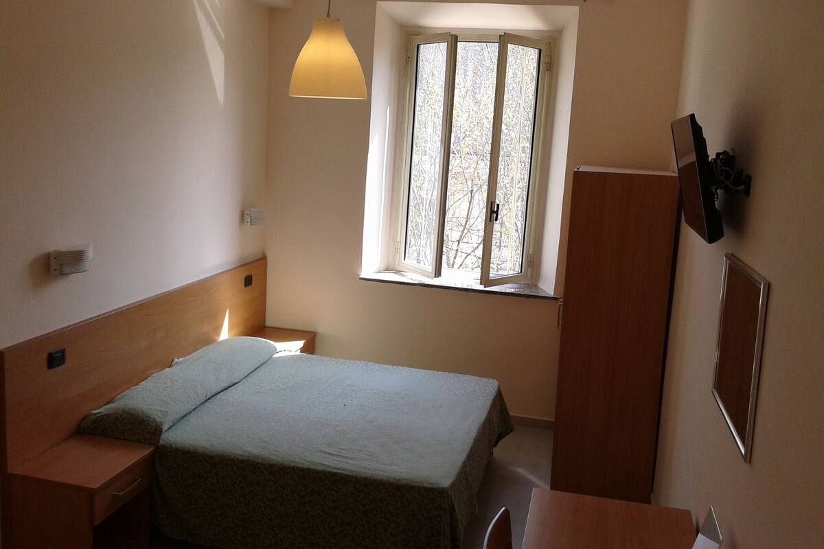 Un dormitorio pequeño y bien iluminado con una cama individual, una mesita de noche, un armario y una ventana con vistas a árboles.