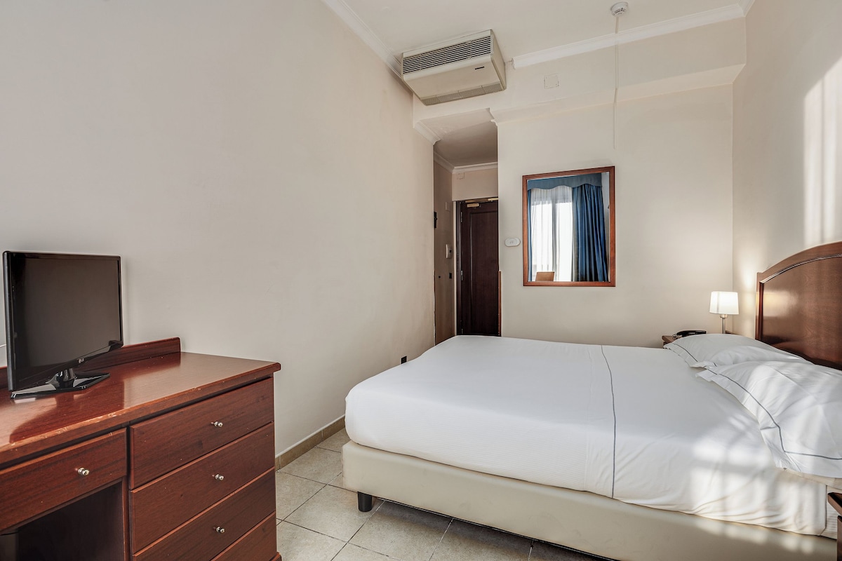 A hotel room with a bed, a TV on a wooden dresser, a bedside lamp, and a window with curtains.