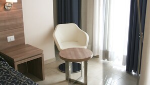 A hotel room with a bed, a small wooden side table, a white chair, and a lamp.