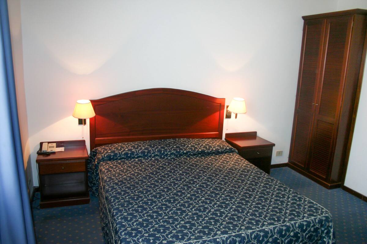 A hotel room with a bed, two bedside tables with lamps, a wardrobe, and a telephone on the left side.
