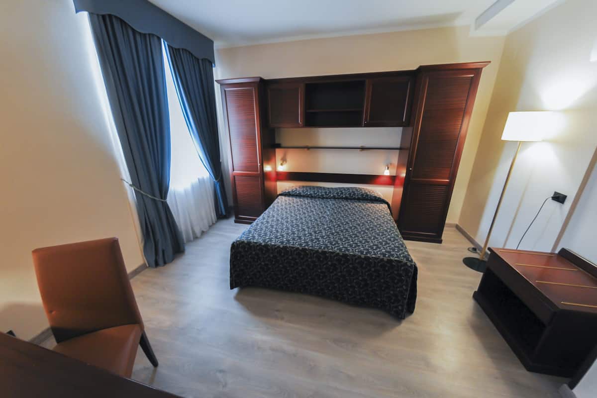 A hotel room with a bed, a desk, a chair, a wardrobe, and a lamp.