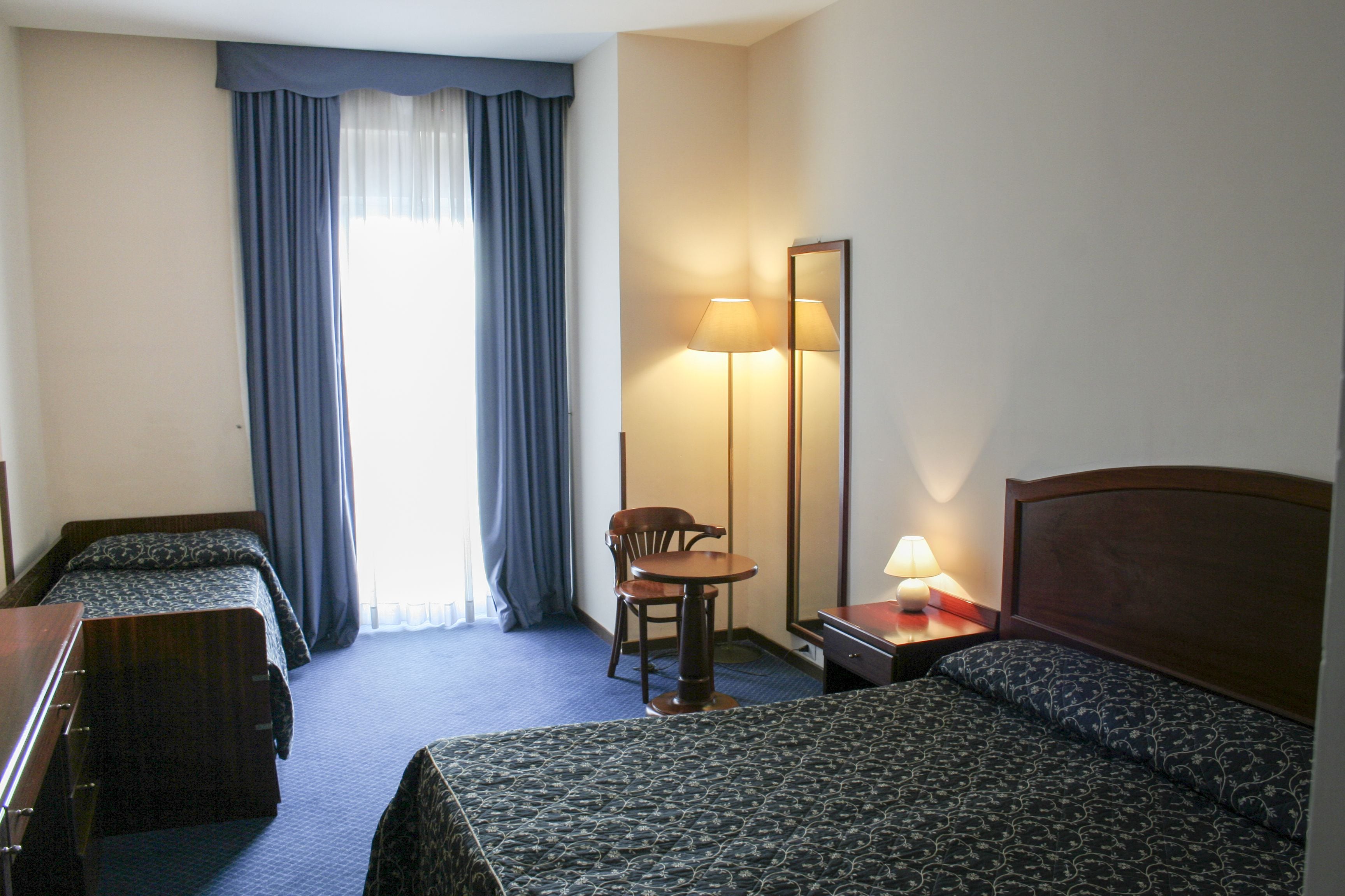 A hotel room with two single beds, a desk with a chair, a standing lamp, and a large mirror.