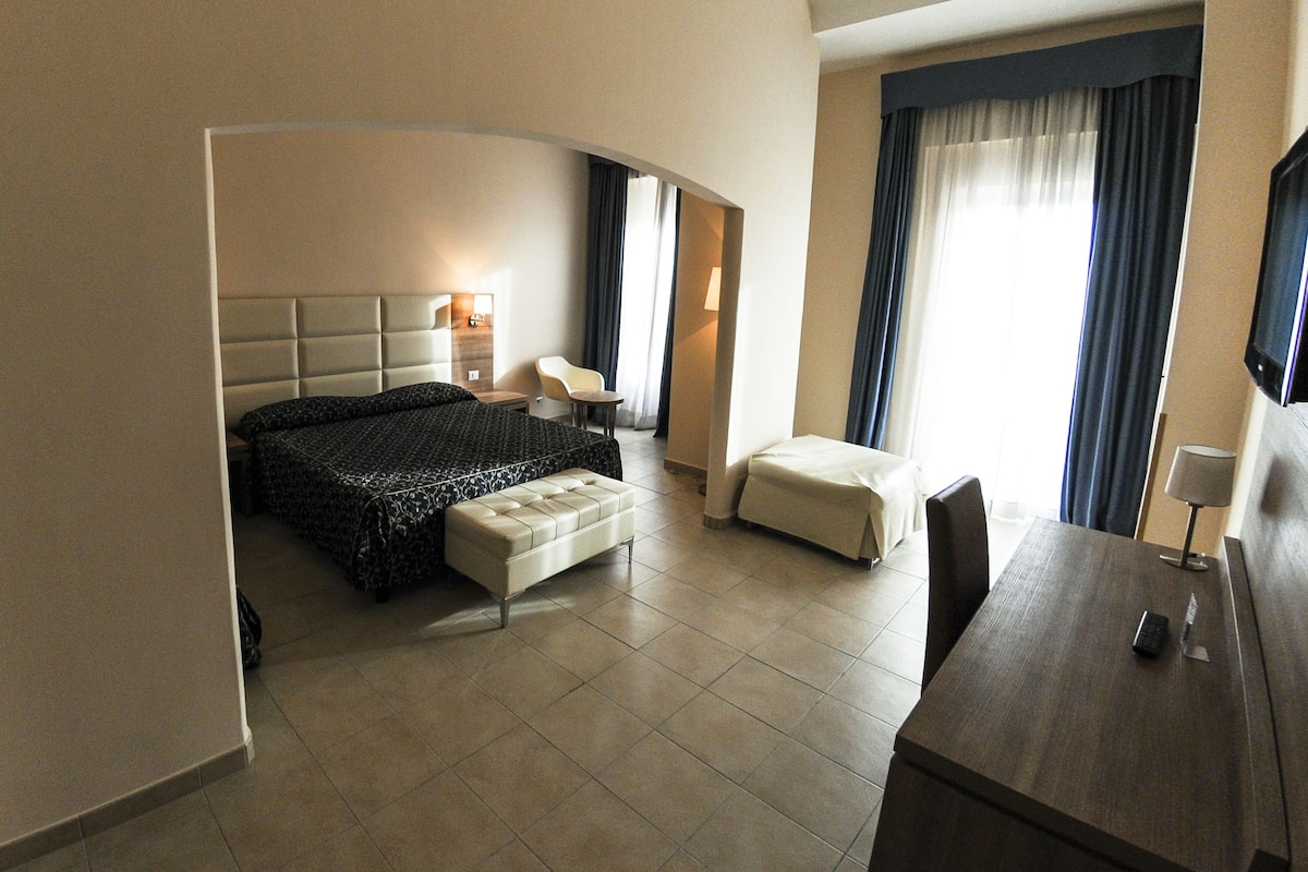 A hotel room with a bed, a small table, a chair, a TV, and a window with curtains.