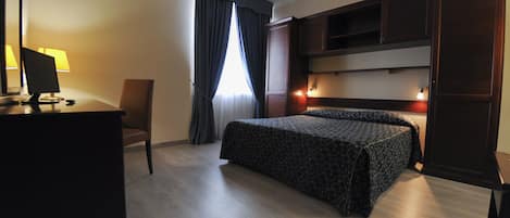 A hotel room with a bed, a desk with a computer, a chair, and a window with curtains.