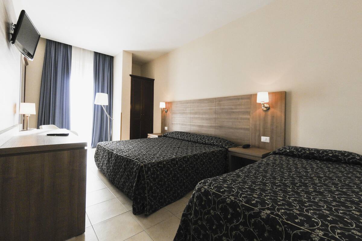 A hotel room with two beds, a desk, a TV, and a window with curtains.