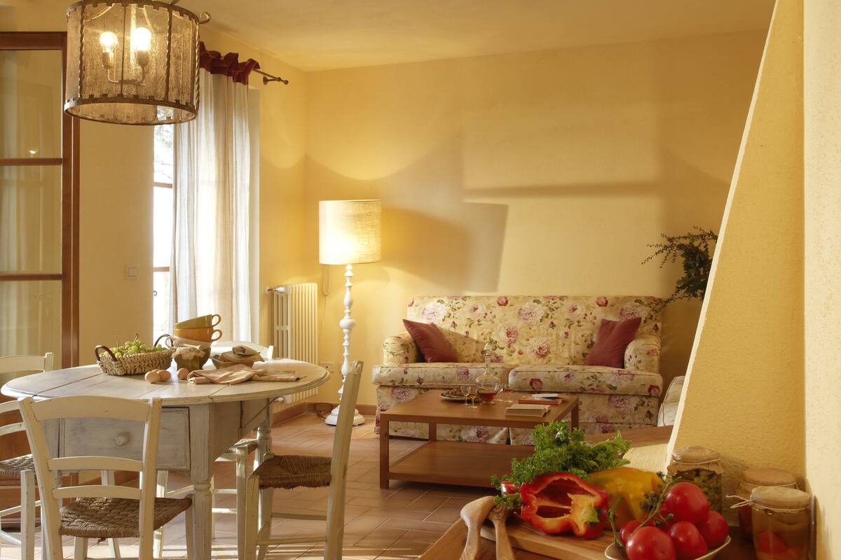 A cozy living room with a floral sofa, a dining table with chairs, and a variety of fruits and vegetables on the table.
