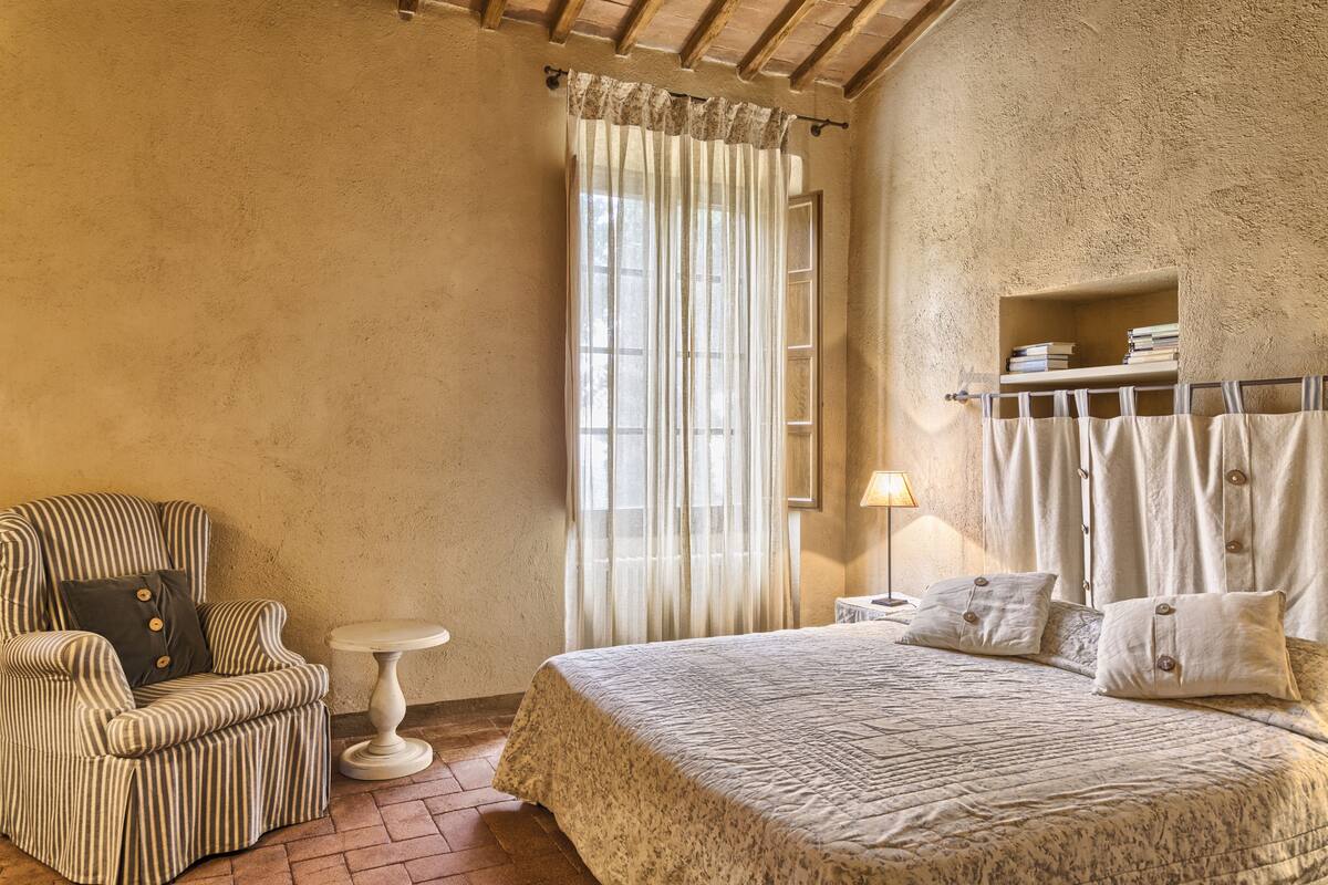 A cozy bedroom with a bed, a striped armchair, a small table, and a shelf with books.