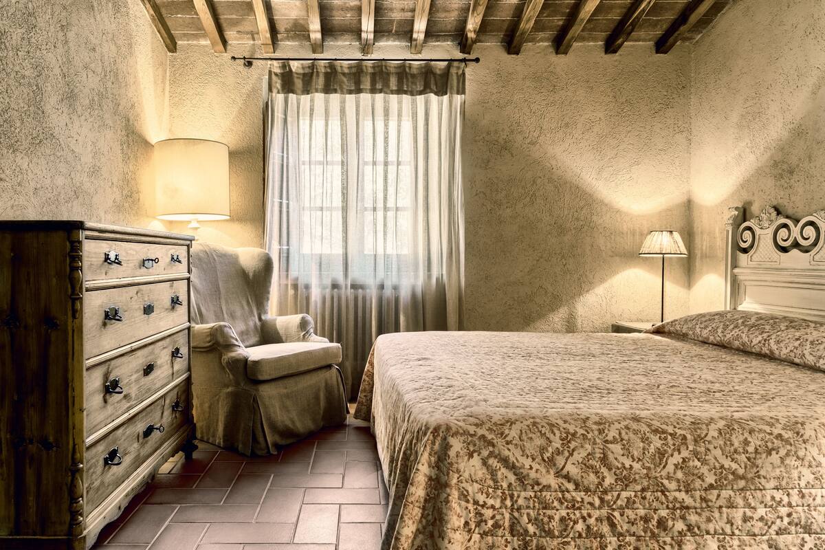 A bedroom with a wooden dresser, a chair, a bed with a floral bedspread, and a window with sheer curtains.