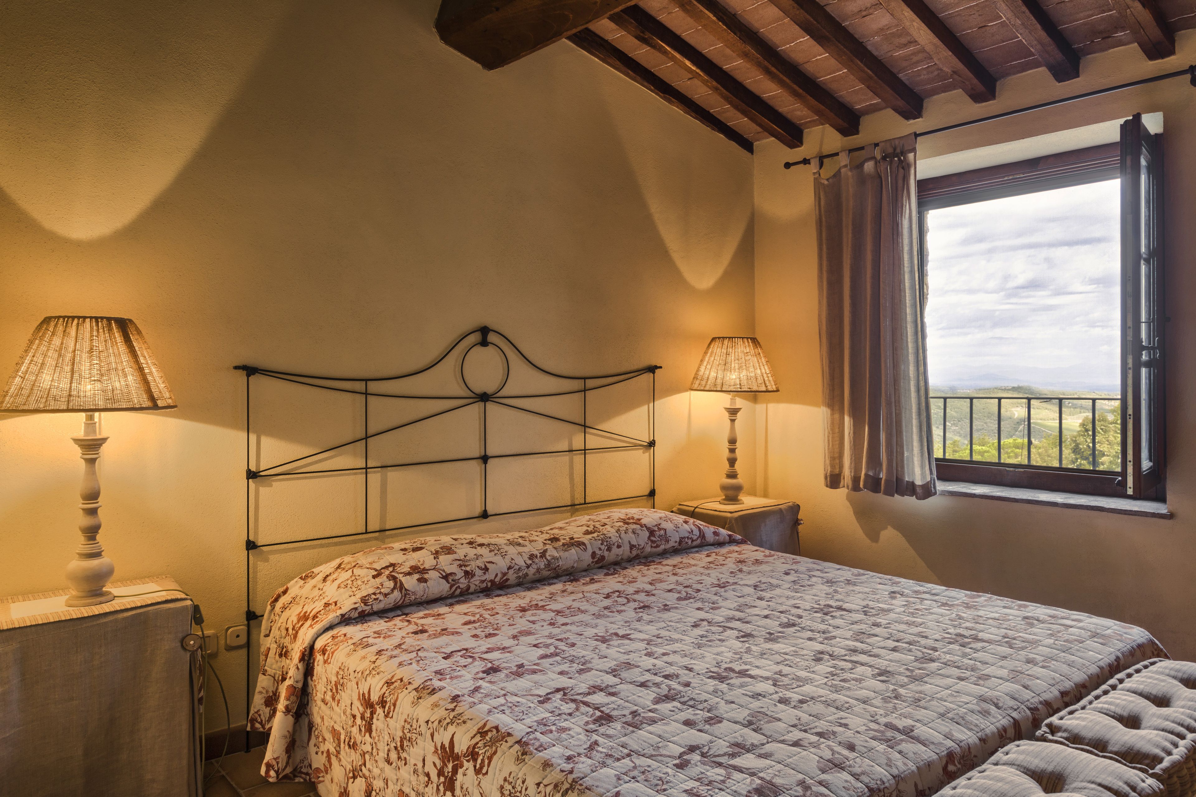 A bedroom with a bed, two bedside lamps, a window with curtains, and a view of the countryside.