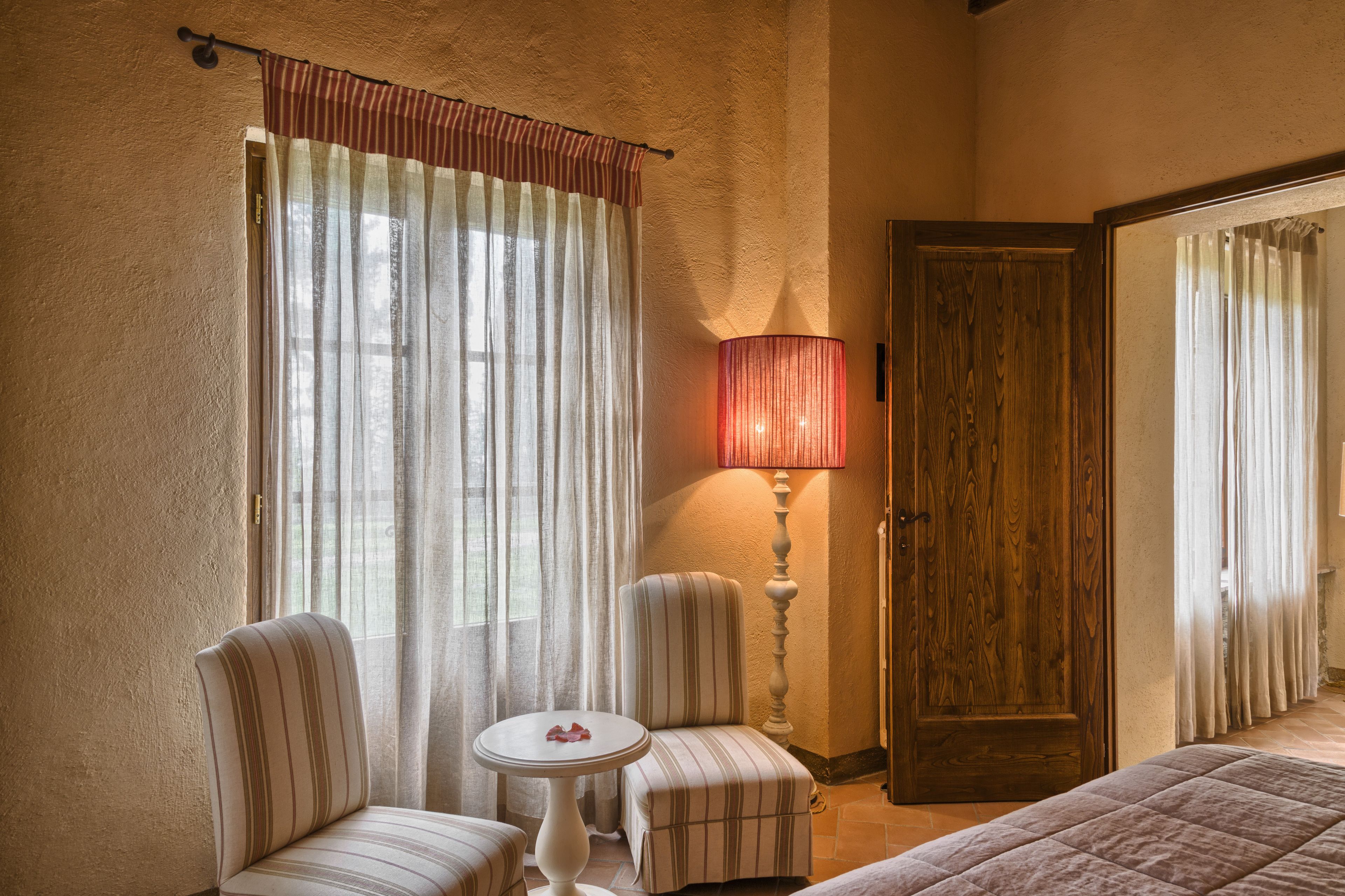 A hotel room with a bed, two striped armchairs, a small round table, a lamp, and a wooden door.