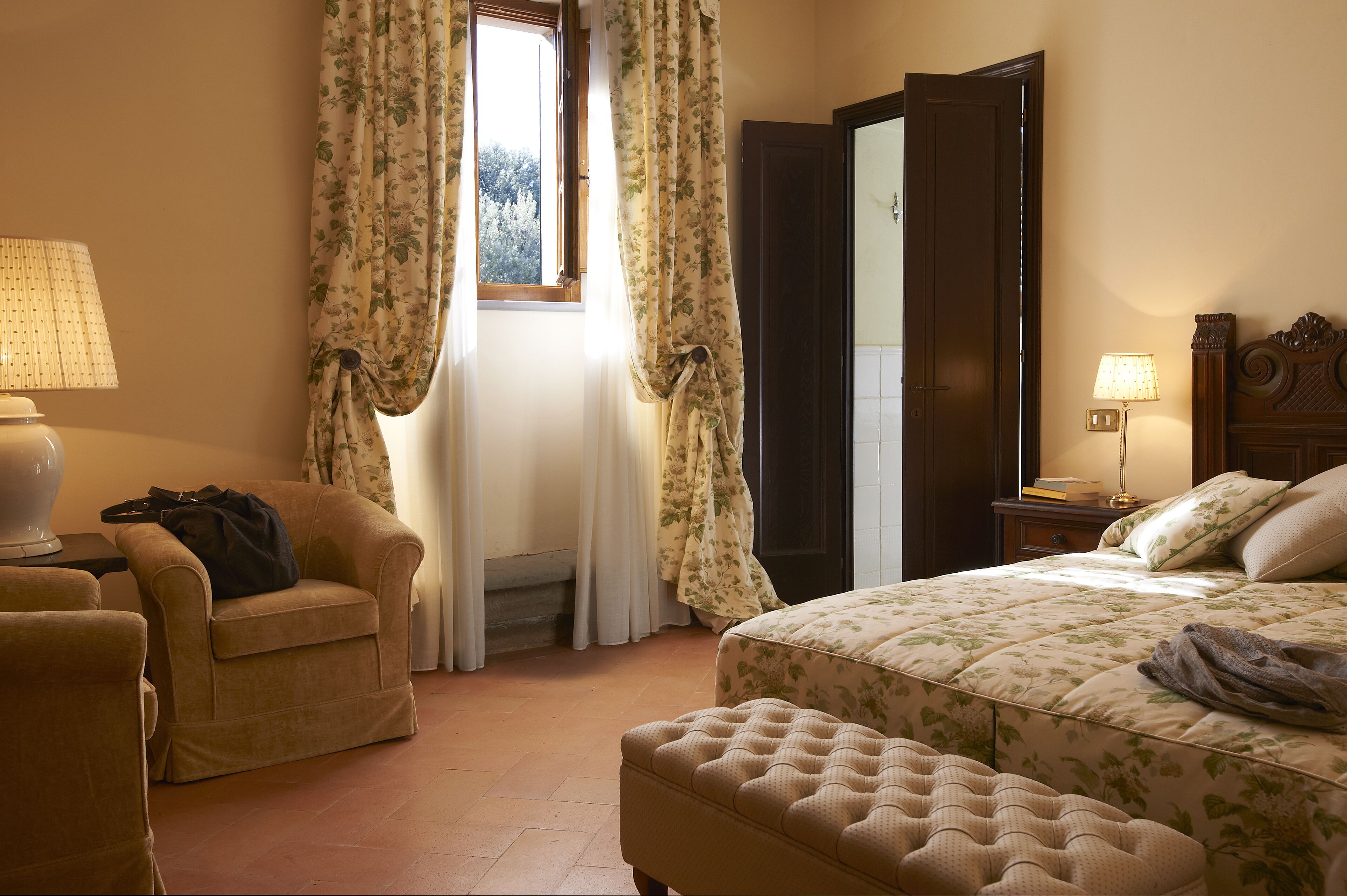 A bedroom with a bed, a chair, a nightstand, a lamp, and a window with curtains.
