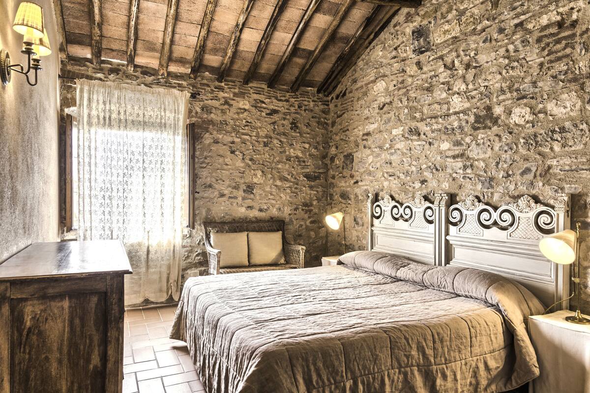 A rustic bedroom with a stone wall, a wooden bed frame, a bedside table, and a lamp.