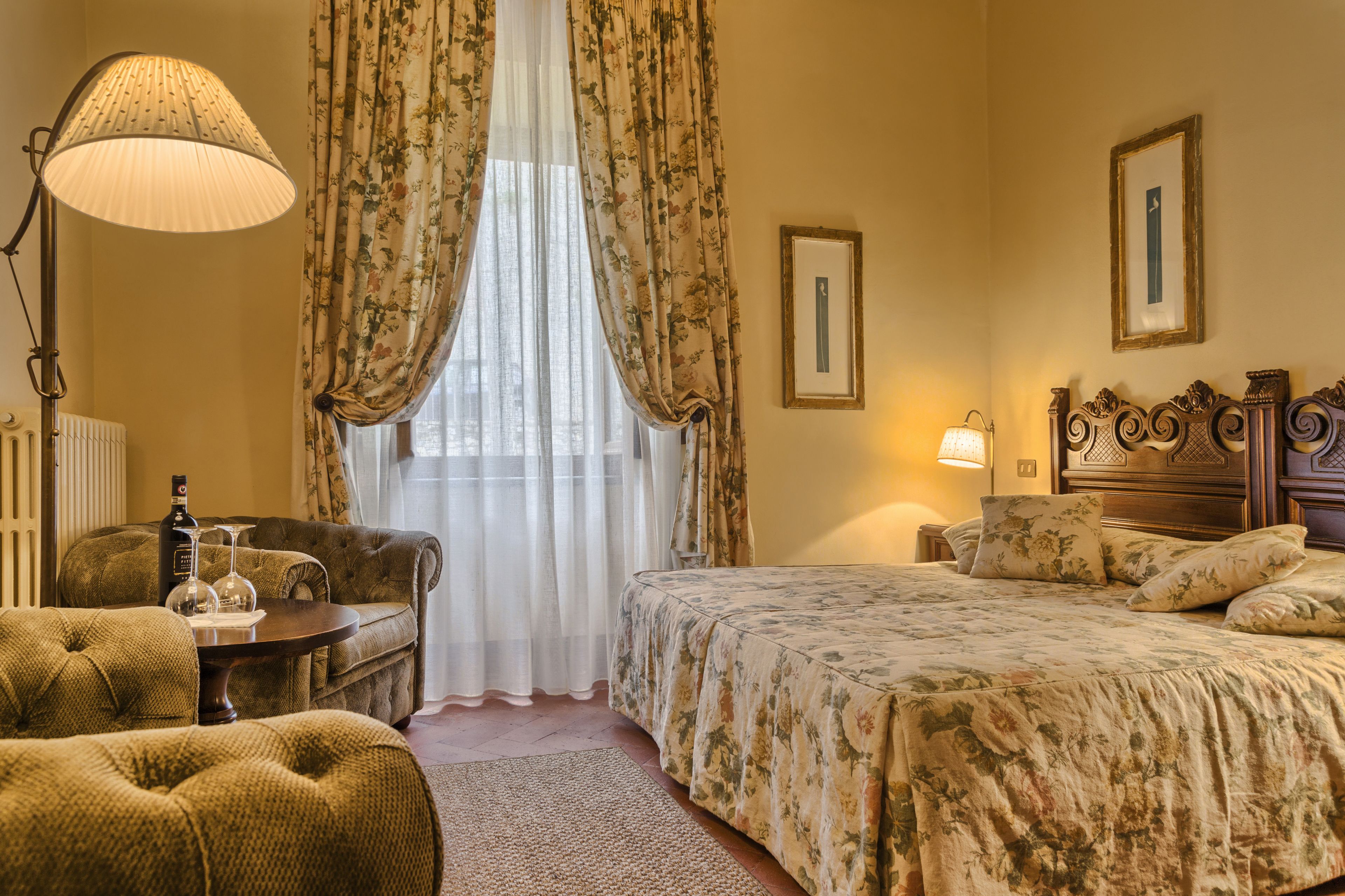 A hotel room with a bed, a sofa, a table with a bottle and glass, a lamp, and curtains.