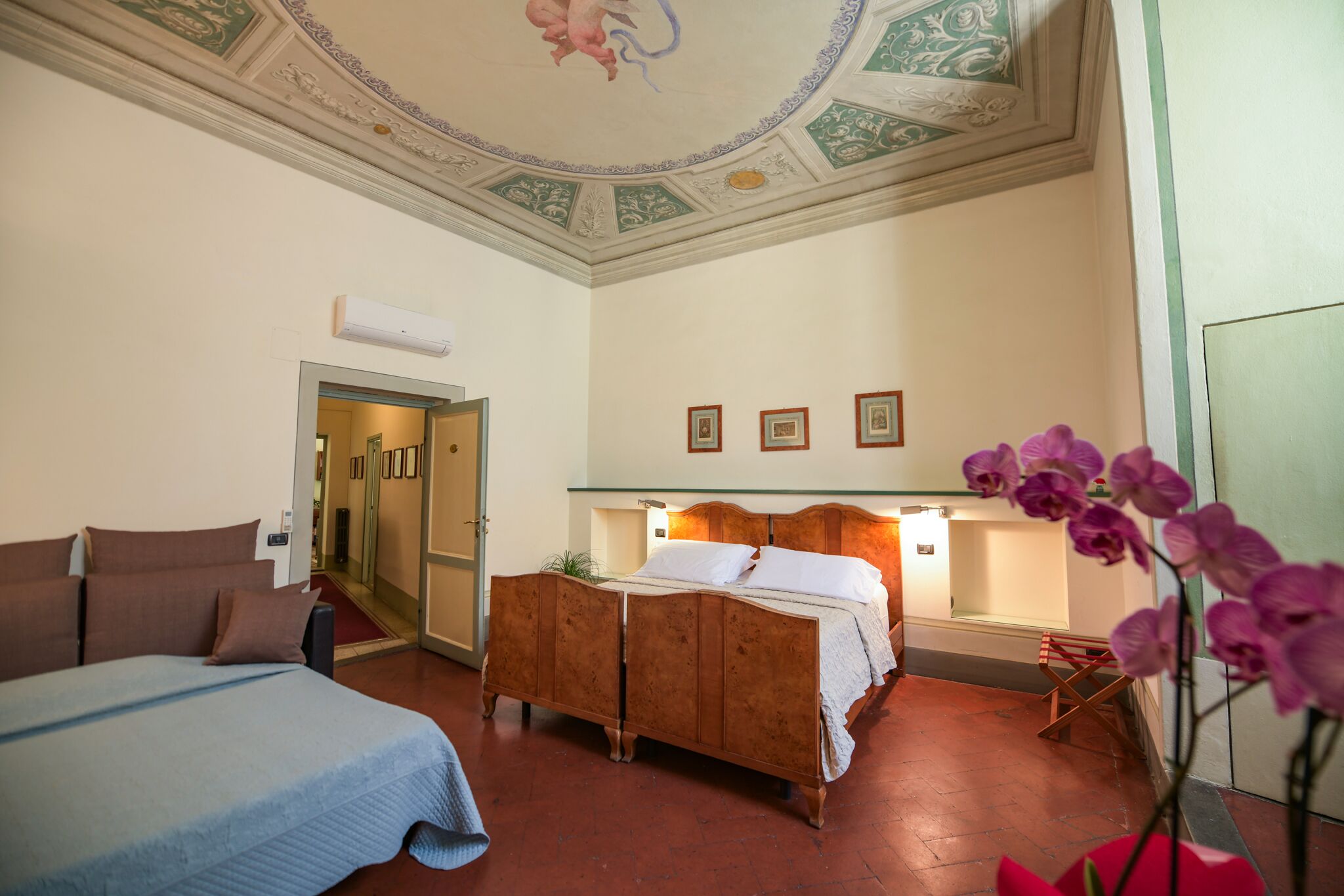 Habitación de hotel con una cama grande, una cama más pequeña, un armario de madera, un escritorio con una silla, un espejo y un techo con diseños decorativos.