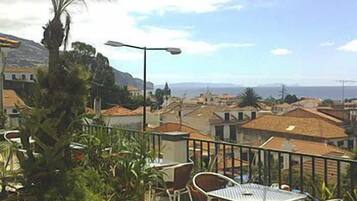 Un balcon avec une table et des chaises, donnant sur une ville côtière avec ses bâtiments et un ciel dégagé.