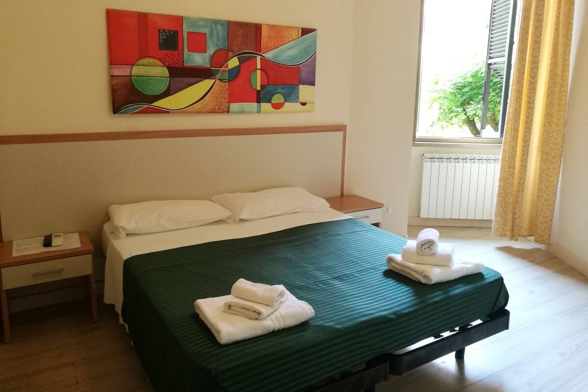 Hotellihuone, jossa on sänky, yöpöytä, taulu ja ikkuna, jossa on verhot.