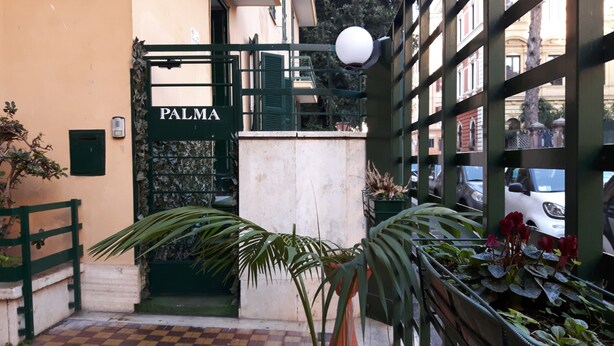 A gated entrance labeled "PALMA" with a checkered floor pattern, potted plants, and a street view through the gate. - Palma Residence (Rome)