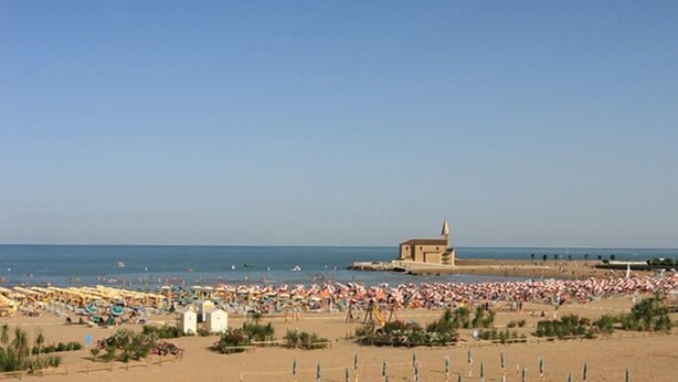 A beach with numerous umbrellas and people. There is a building with a pointed roof in the distance.