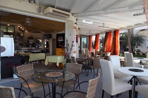 An outdoor cafe with wicker chairs and tables, a kitchen area visible through an open door, and orange curtains. - Aparthotel Gioia (Caorle)