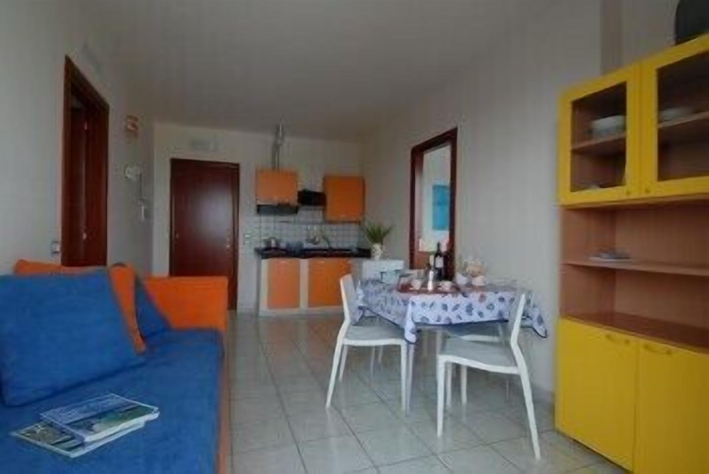 A compact living space with a blue sofa, a dining table with chairs, a kitchen area with orange cabinets, and a yellow cabinet.