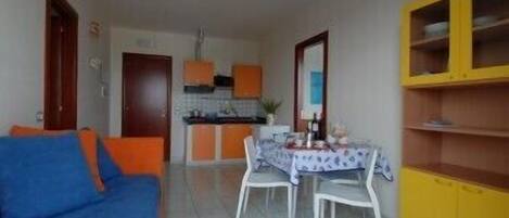 A compact living space with a blue sofa, a dining table with chairs, a kitchen area with orange cabinets, and a yellow cabinet.