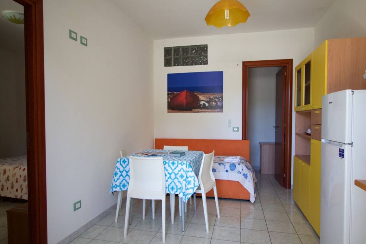 Une pièce de taille modeste comprenant une table à manger, des chaises, un lit, un réfrigérateur et une lampe suspendue jaune.