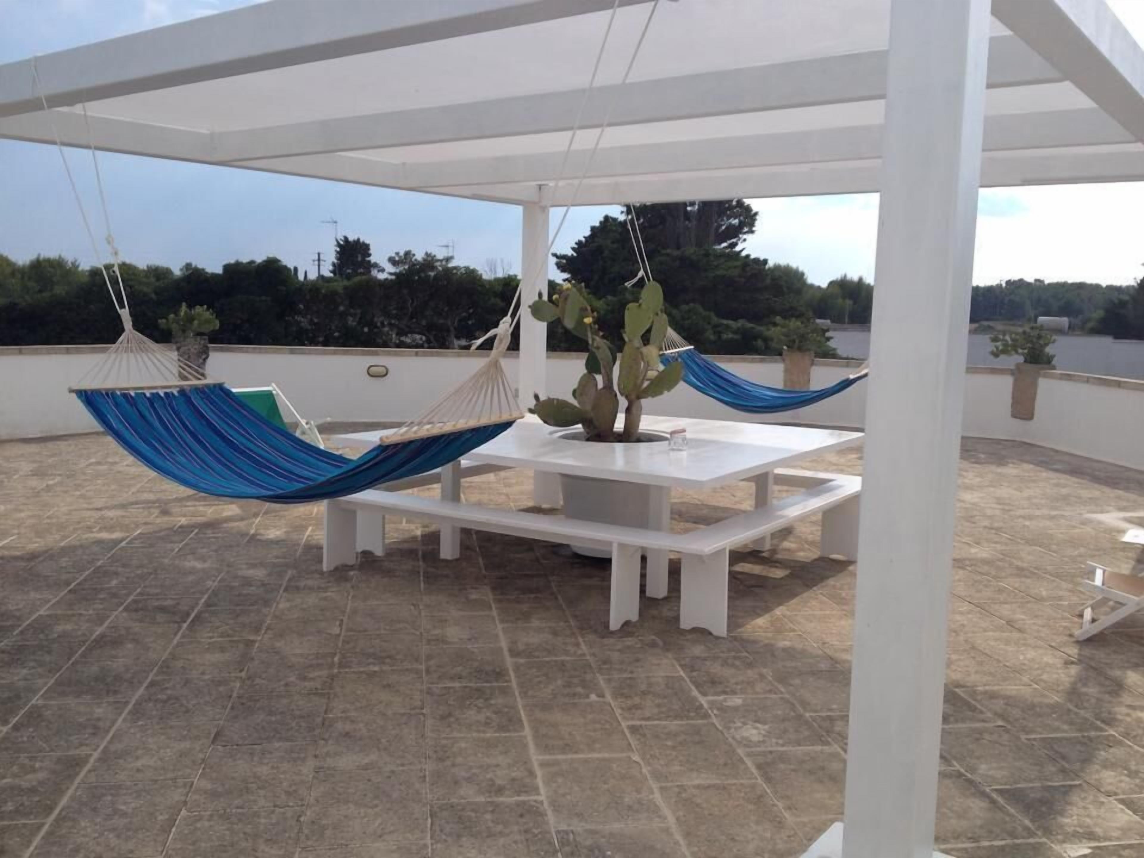 Un patio couvert avec une table et des bancs blancs, deux hamacs bleus et un cactus.