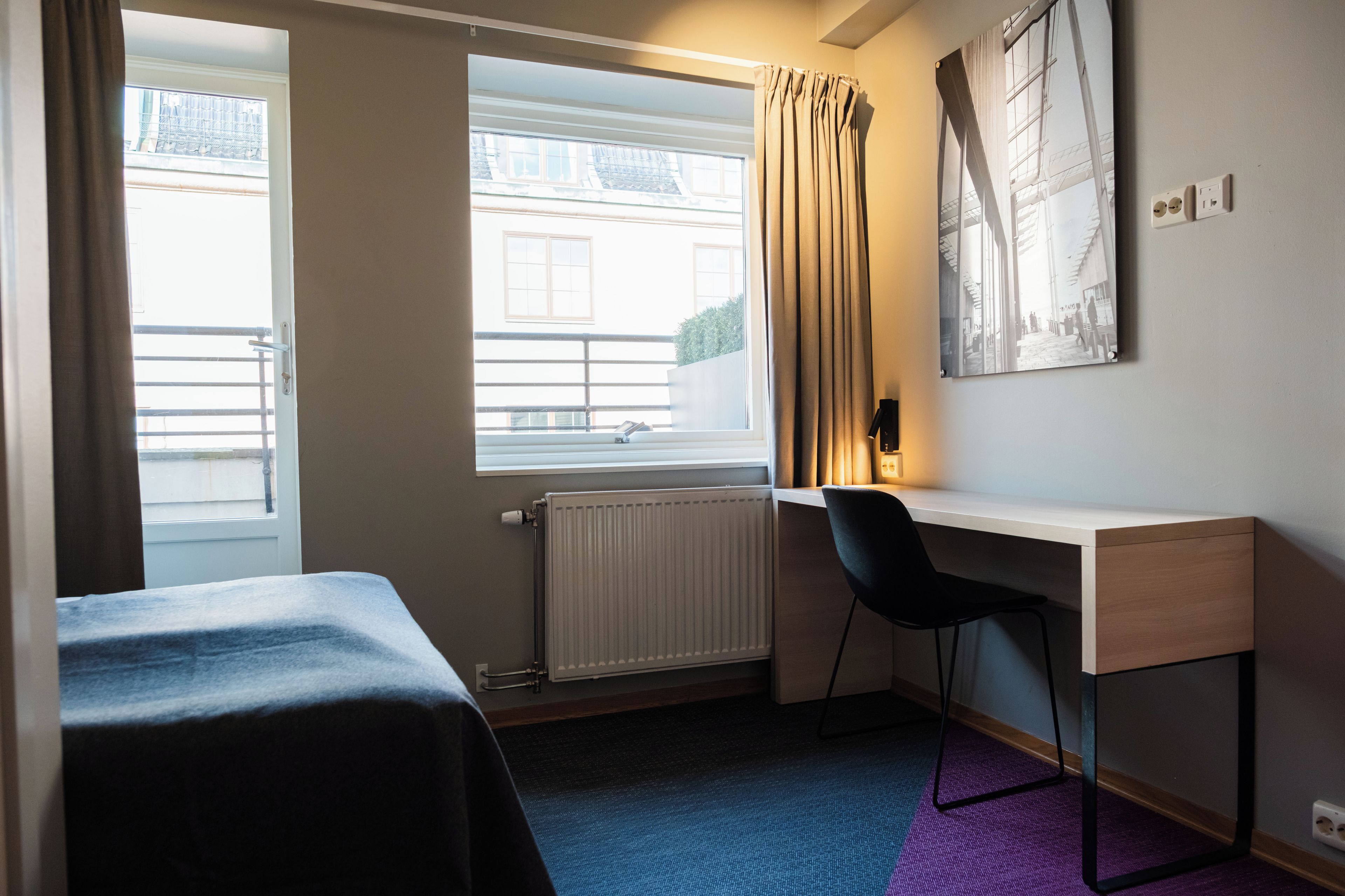 A hotel room with a bed, a desk, a chair, a radiator, a window with curtains, and a wall-mounted picture.