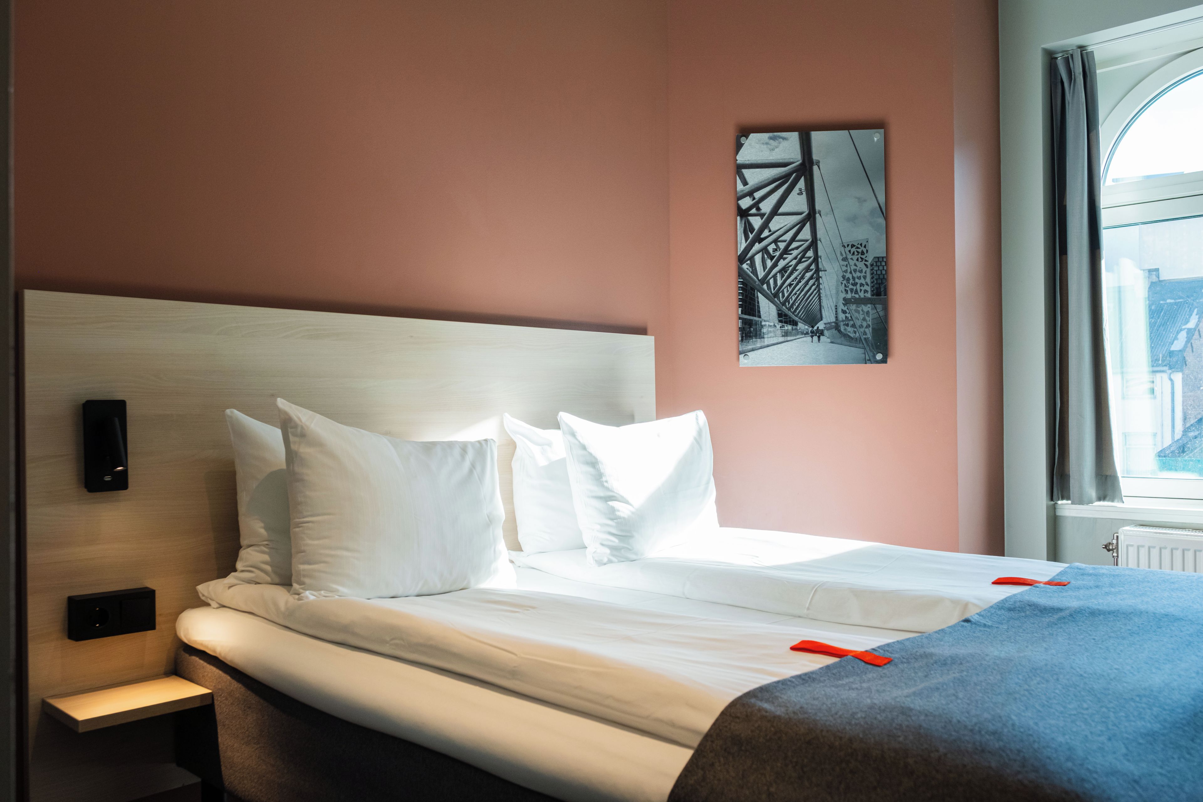 Habitación de hotel con cama, almohadas, mesita de noche, teléfono en la pared y ventana con cortinas.