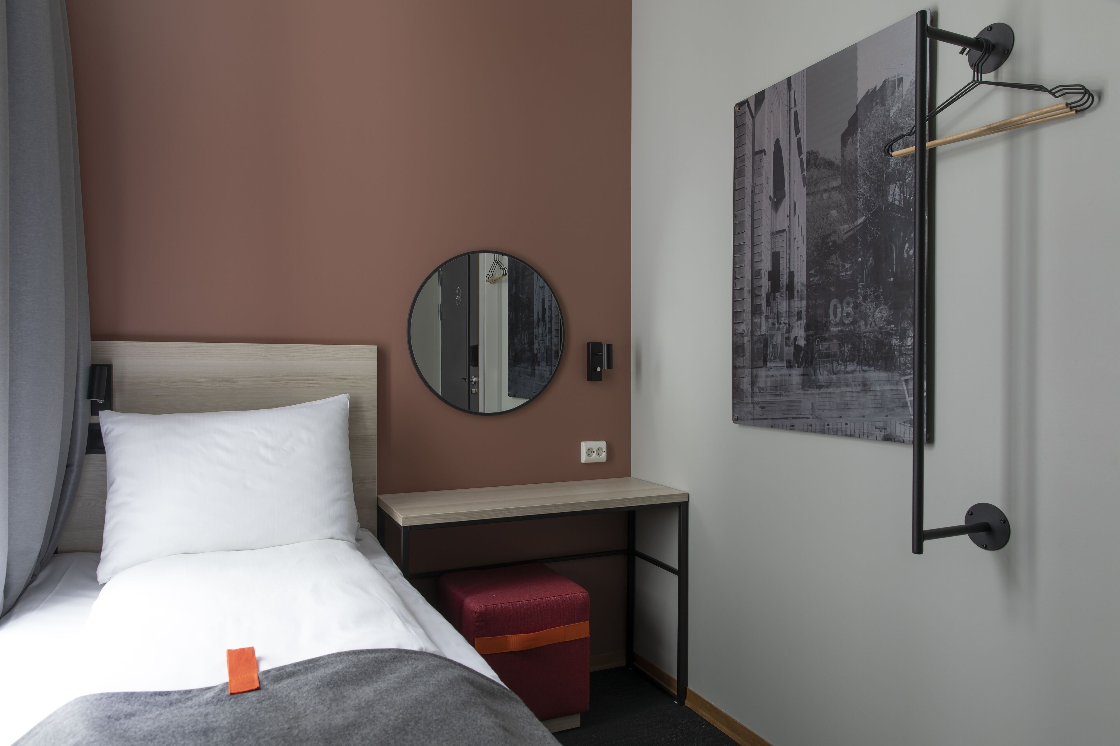 Una habitación de hotel con una cama, un escritorio, un reposapiés rojo, un espejo redondo y una fotografía blanco y negro en la pared.