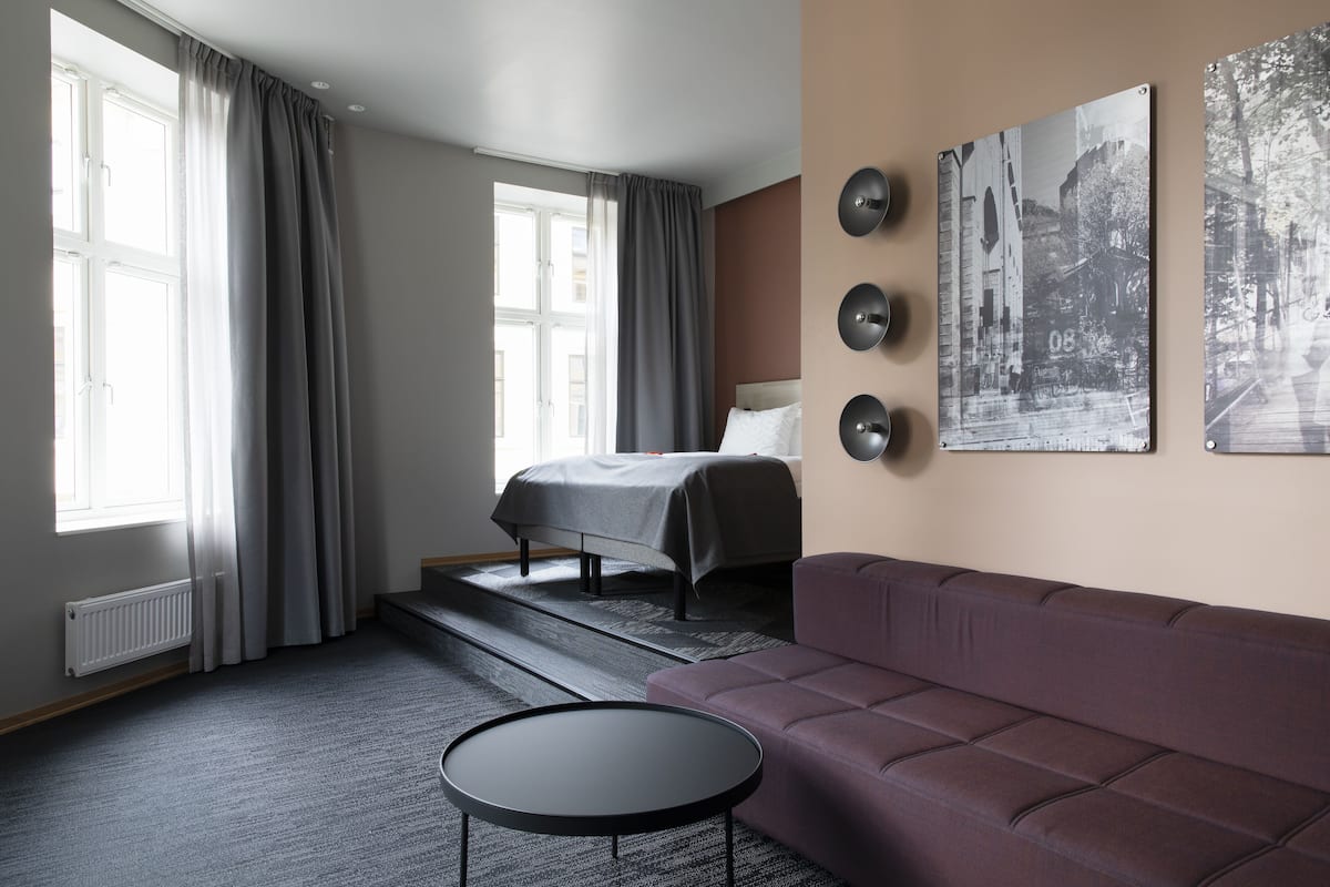 Moderni hotellihuone, jossa on sänky, sohva, pyöreä pöytä ja suuret ikkunat, joissa on verhot.