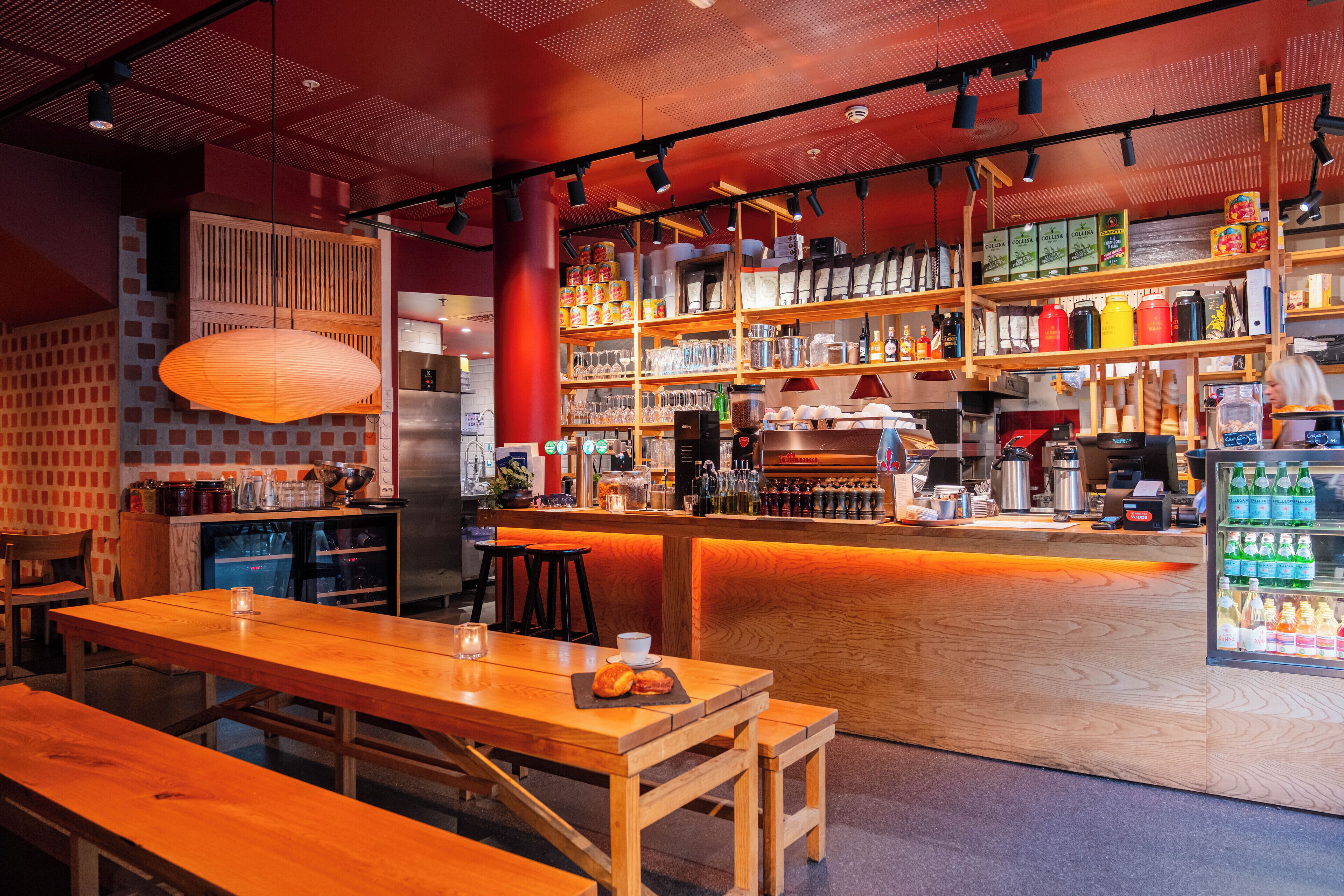 Un café acogedor con mesas y bancos de madera, una zona de bar con estantes llenos de diversos artículos y un expositor refrigerado con bebidas.