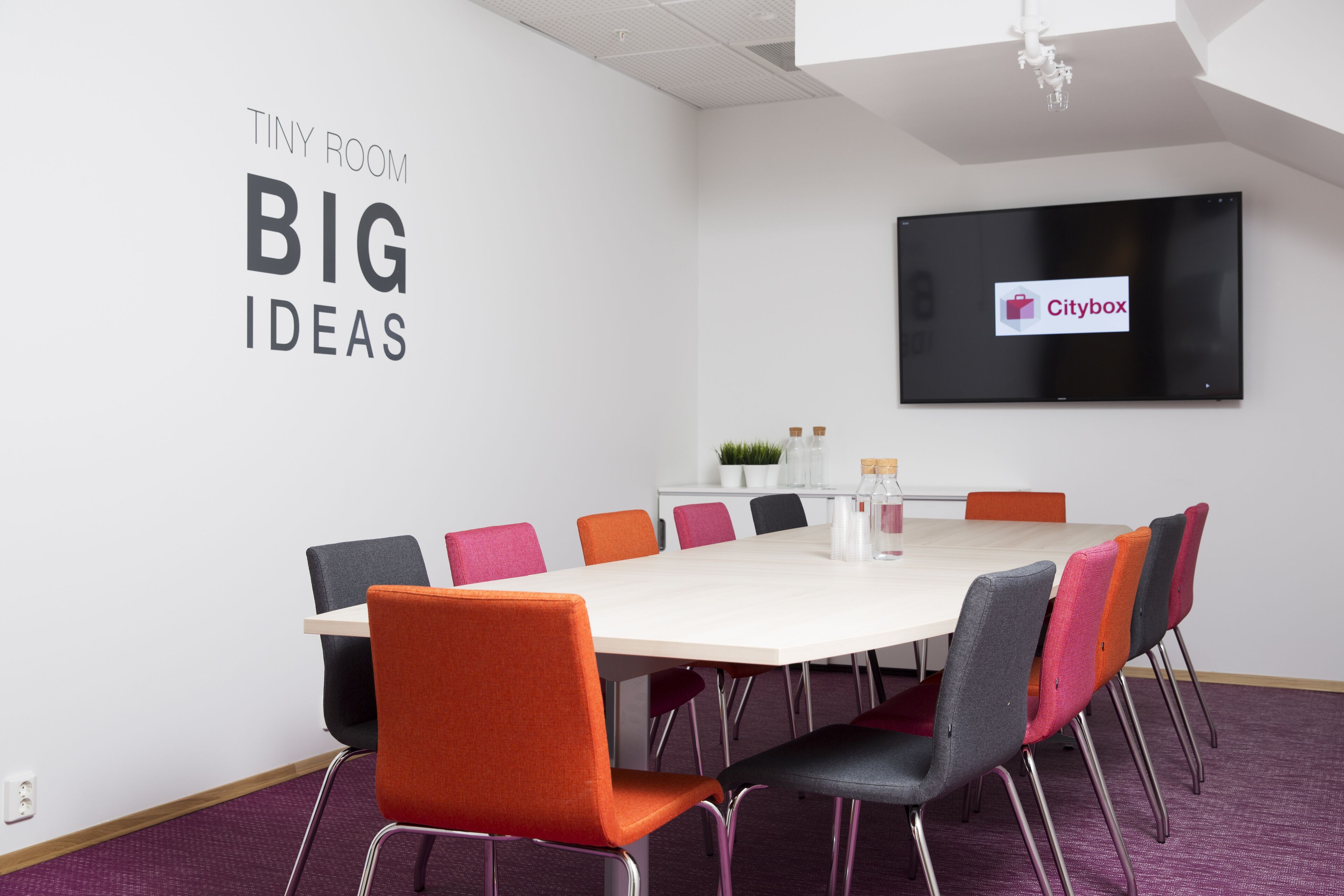 A conference room with a white table, colorful chairs, a mounted TV, and a sign reading "TINY ROOM BIG IDEAS".