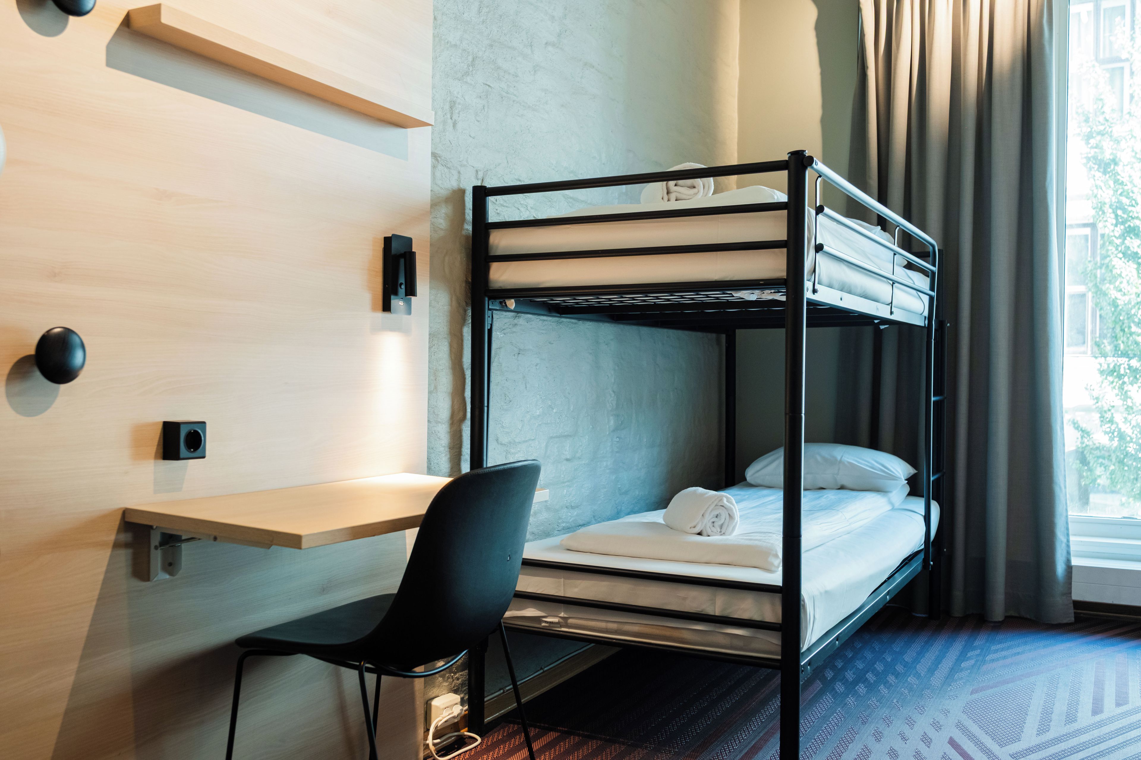 Un lit superposé avec un bureau et une chaise, une lampe fixée au mur et une fenêtre avec des rideaux.