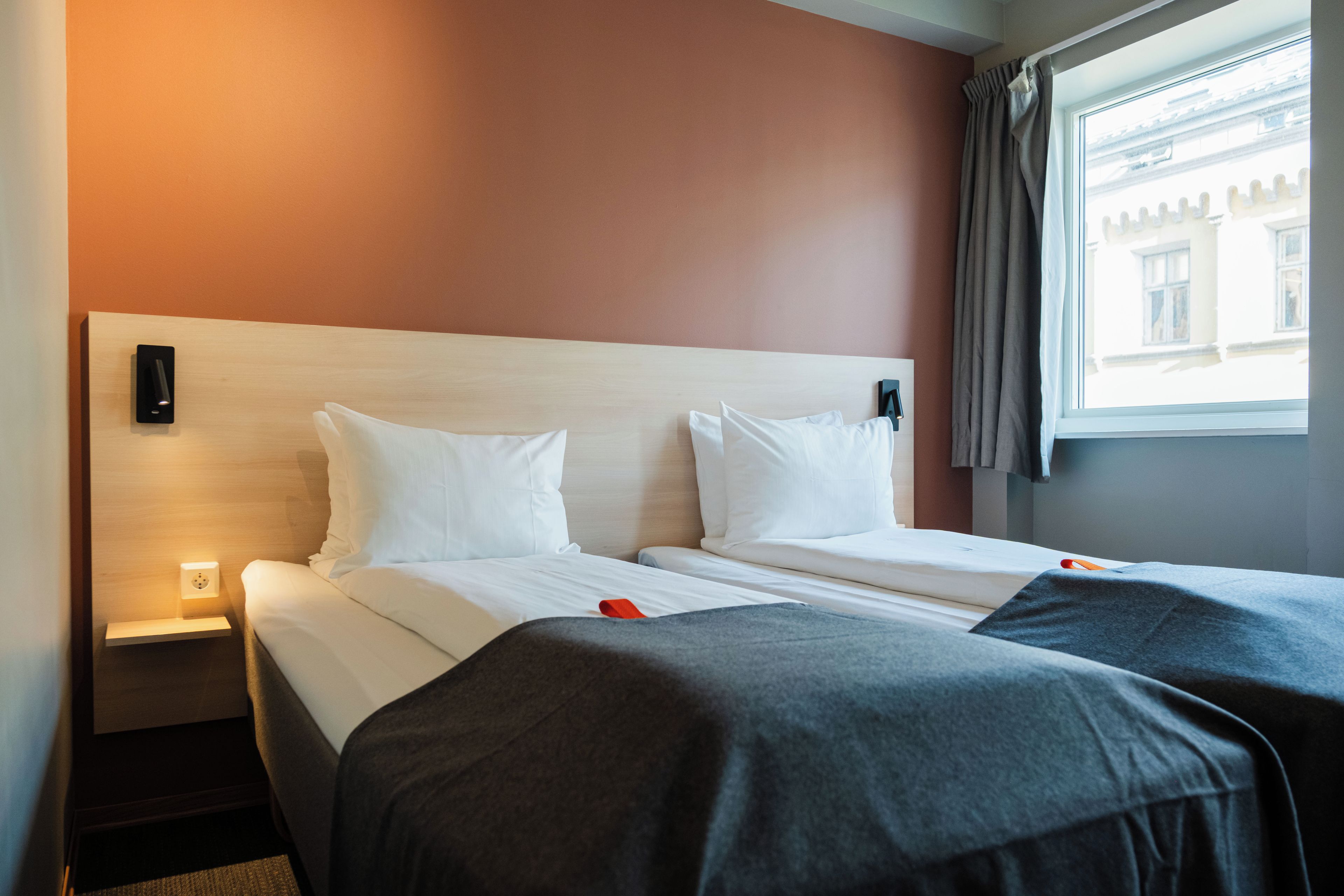 A hotel room with two beds, a nightstand, a wall-mounted light, and a window with curtains.