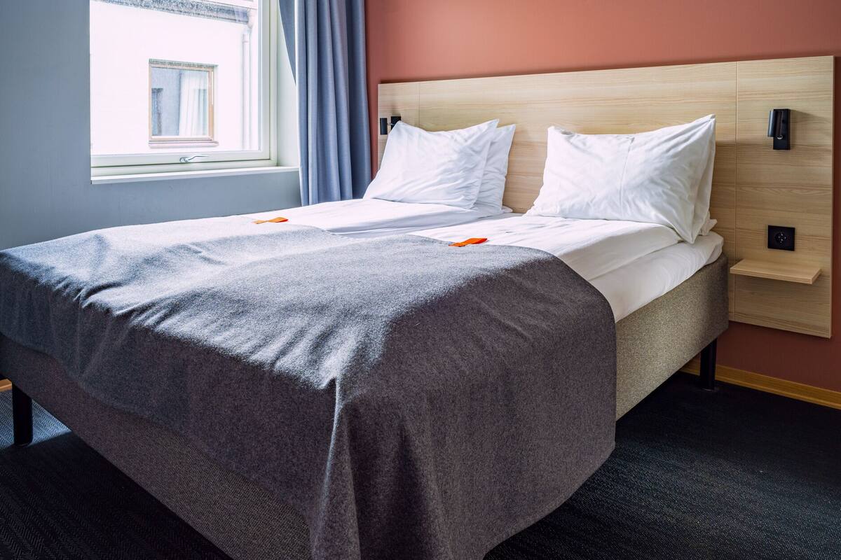 Hotellihuone, jossa on sänky, ikkuna verhoilla, puinen pääty ja ohjauspaneeli seinässä.