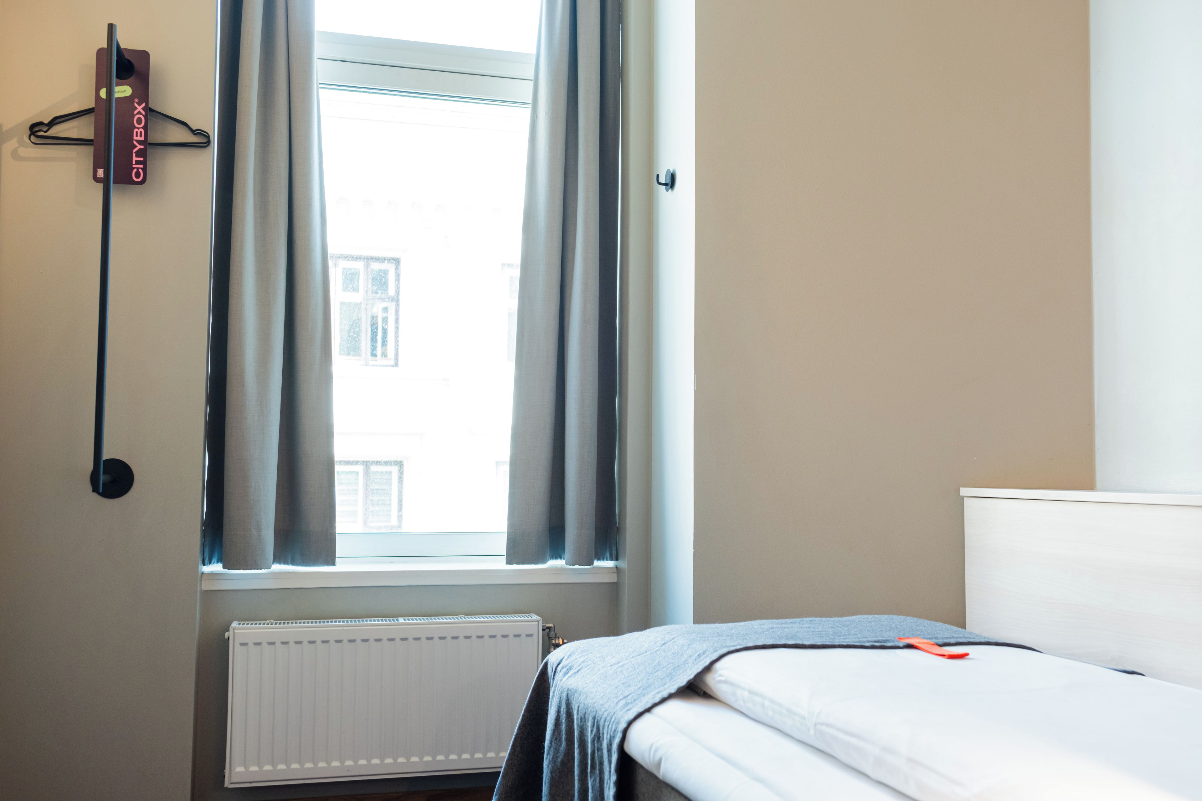 A hotel room with a bed, a radiator, a window with curtains, and a CityBox on the wall.