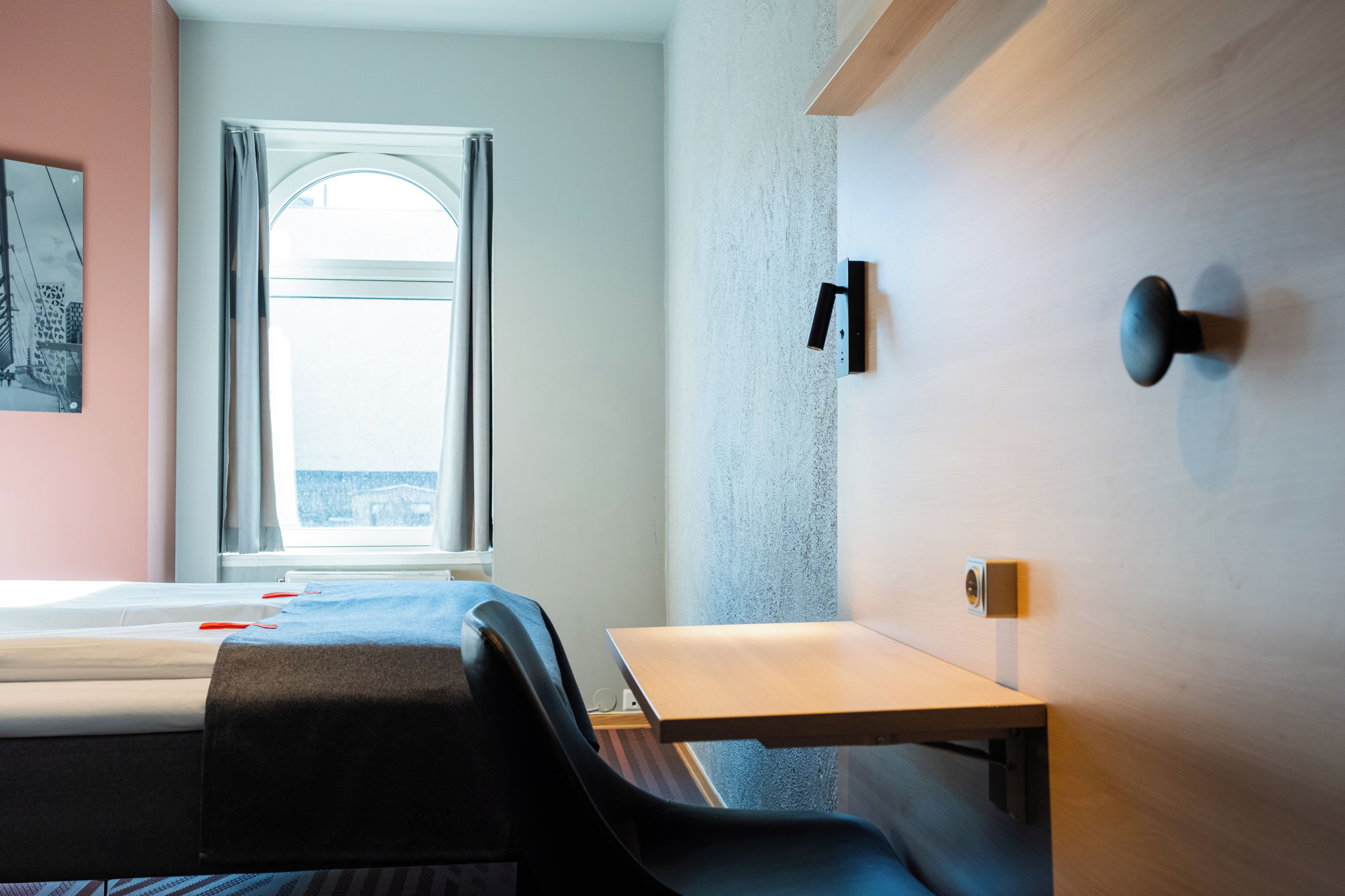 Habitación de hotel con cama, escritorio y silla. Cuenta con una ventana con cortinas, una lámpara de pared y una puerta con manija.