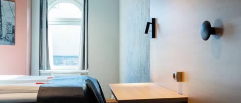 Habitación de hotel con cama, escritorio y silla. Cuenta con una ventana con cortinas, una lámpara de pared y una puerta con manija.
