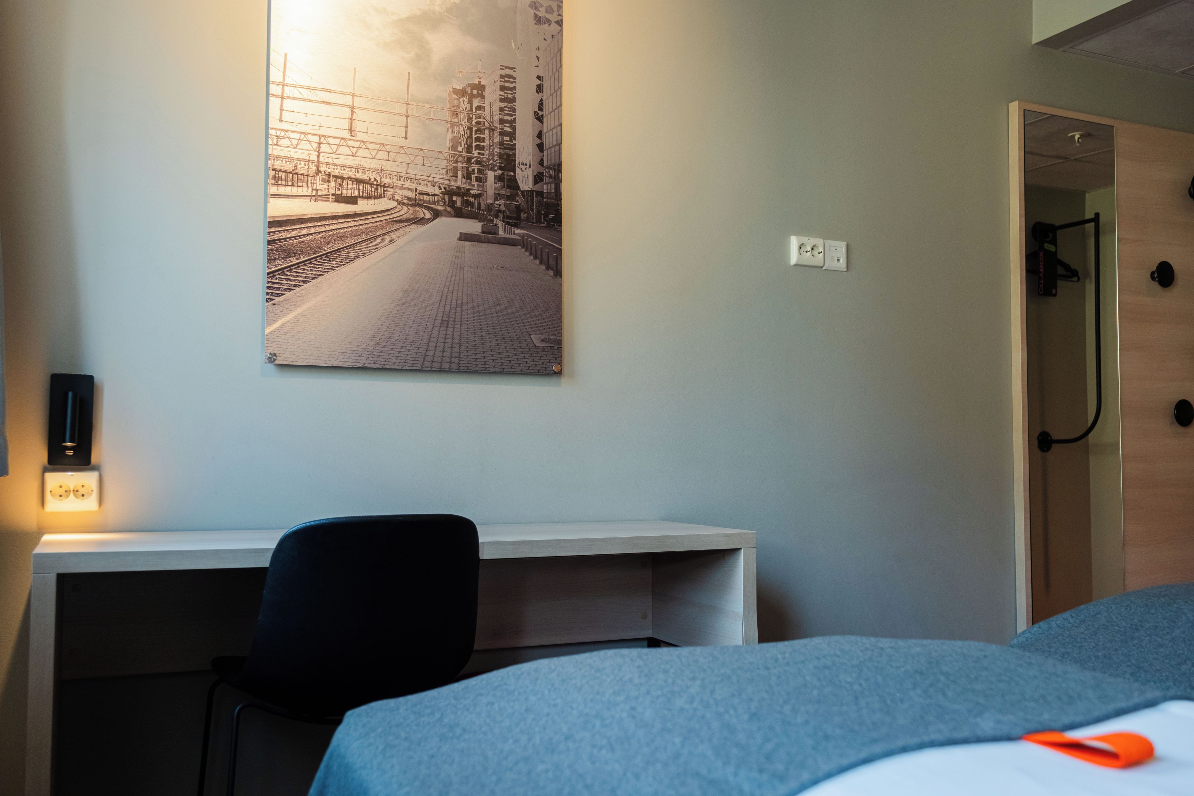 Habitación de hotel con cama, escritorio, silla, un cuadro en la pared y una puerta.
