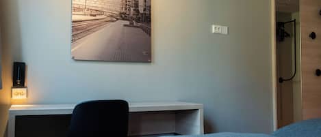 Habitación de hotel con cama, escritorio, silla, un cuadro en la pared y una puerta.