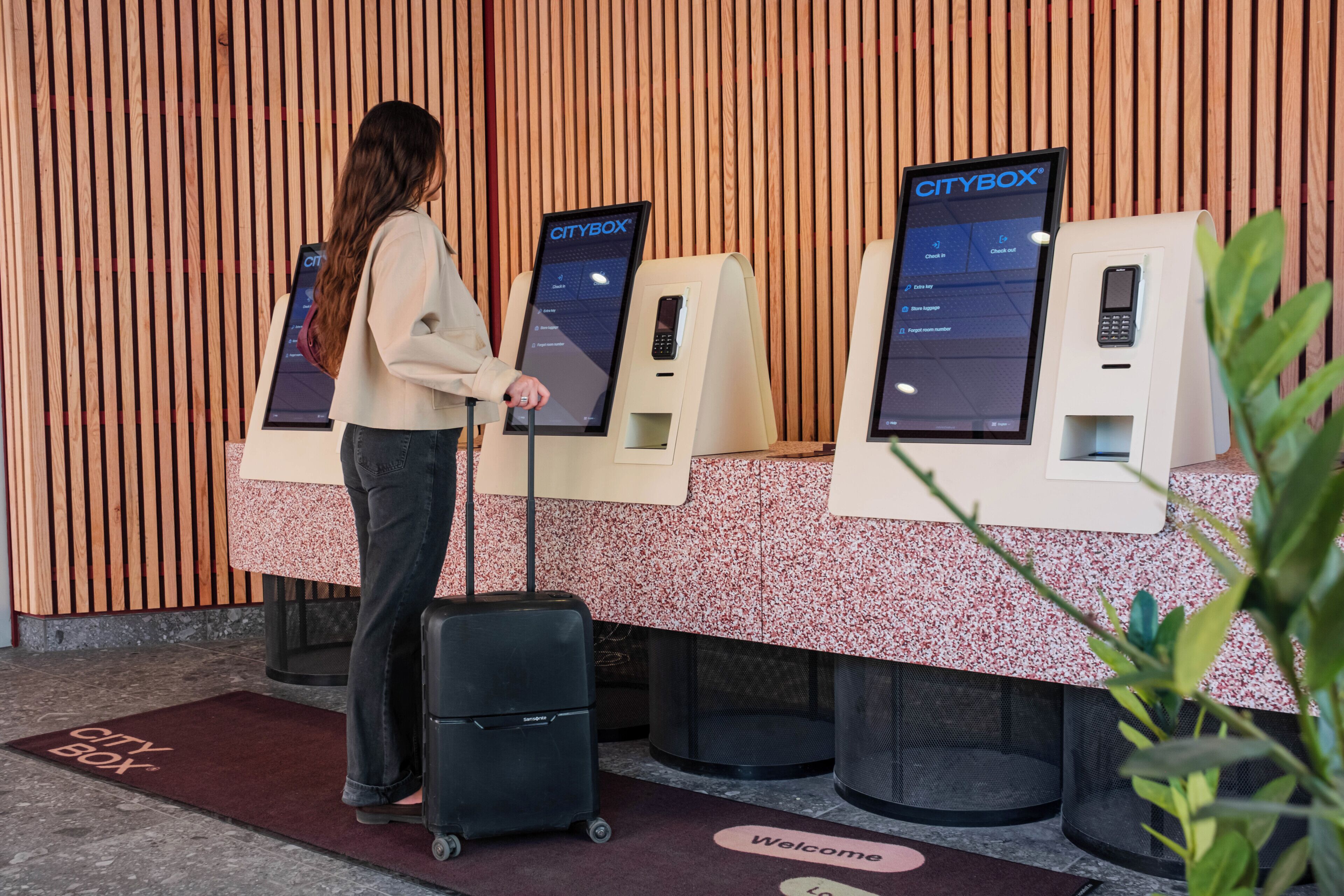A woman at a CityBox self-service kiosk with a suitcase, a welcome mat, and a 'Welcome' sign on the floor.