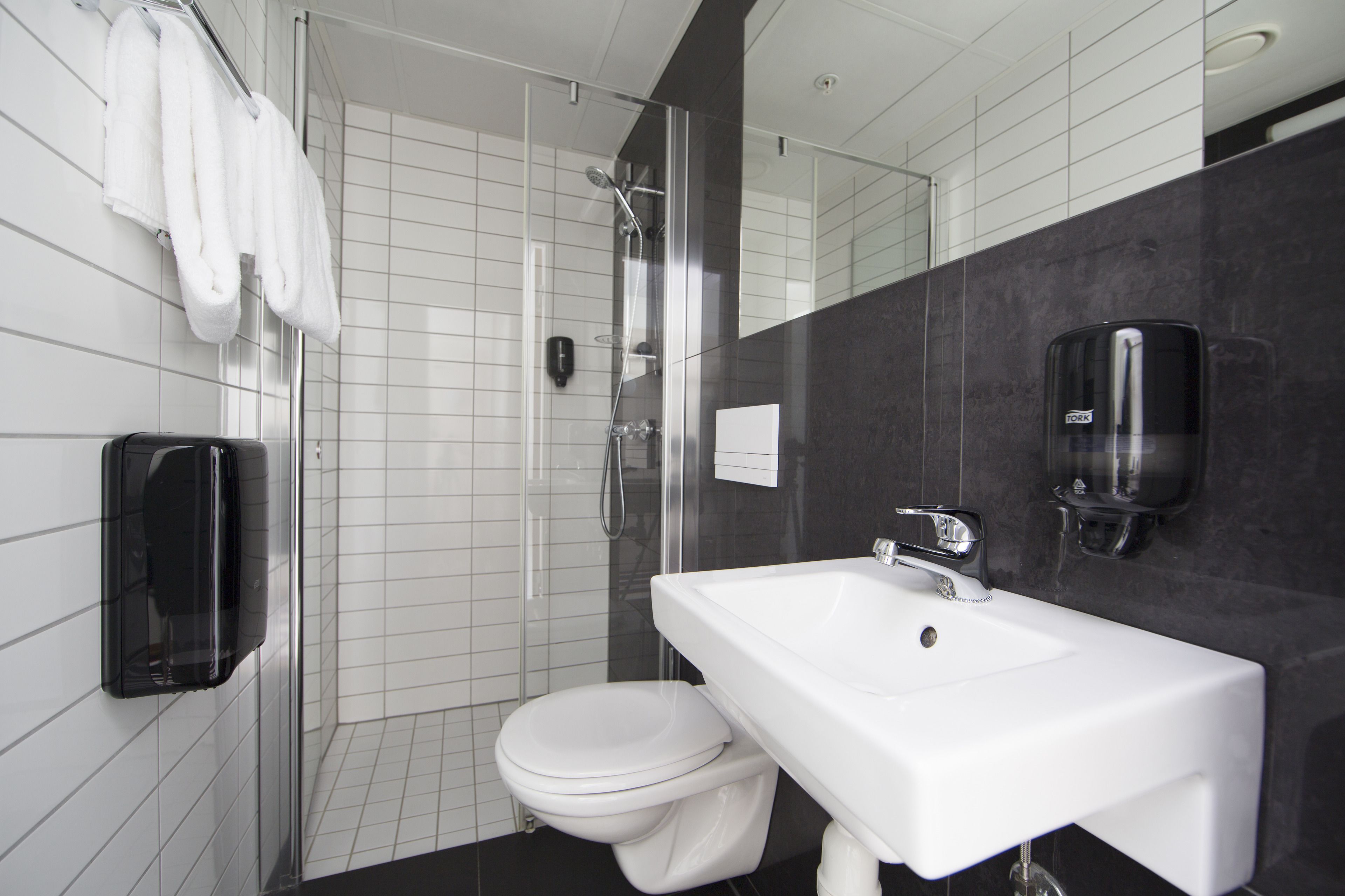 Un baño moderno con un lavamanos blanco, un inodoro, una ducha con mampara de vidrio y un perchero con toallas blancas.