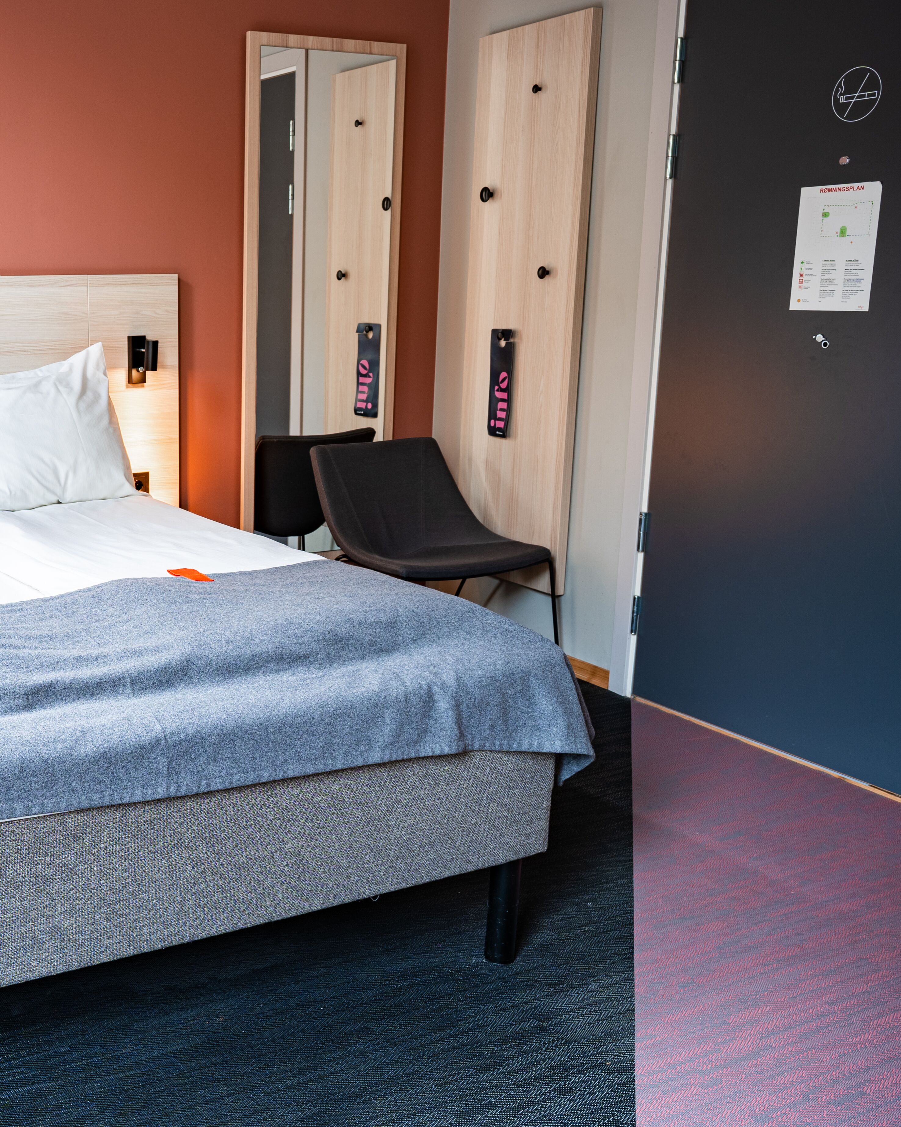 A hotel room with a bed, a chair, a desk, and a door with a sign.