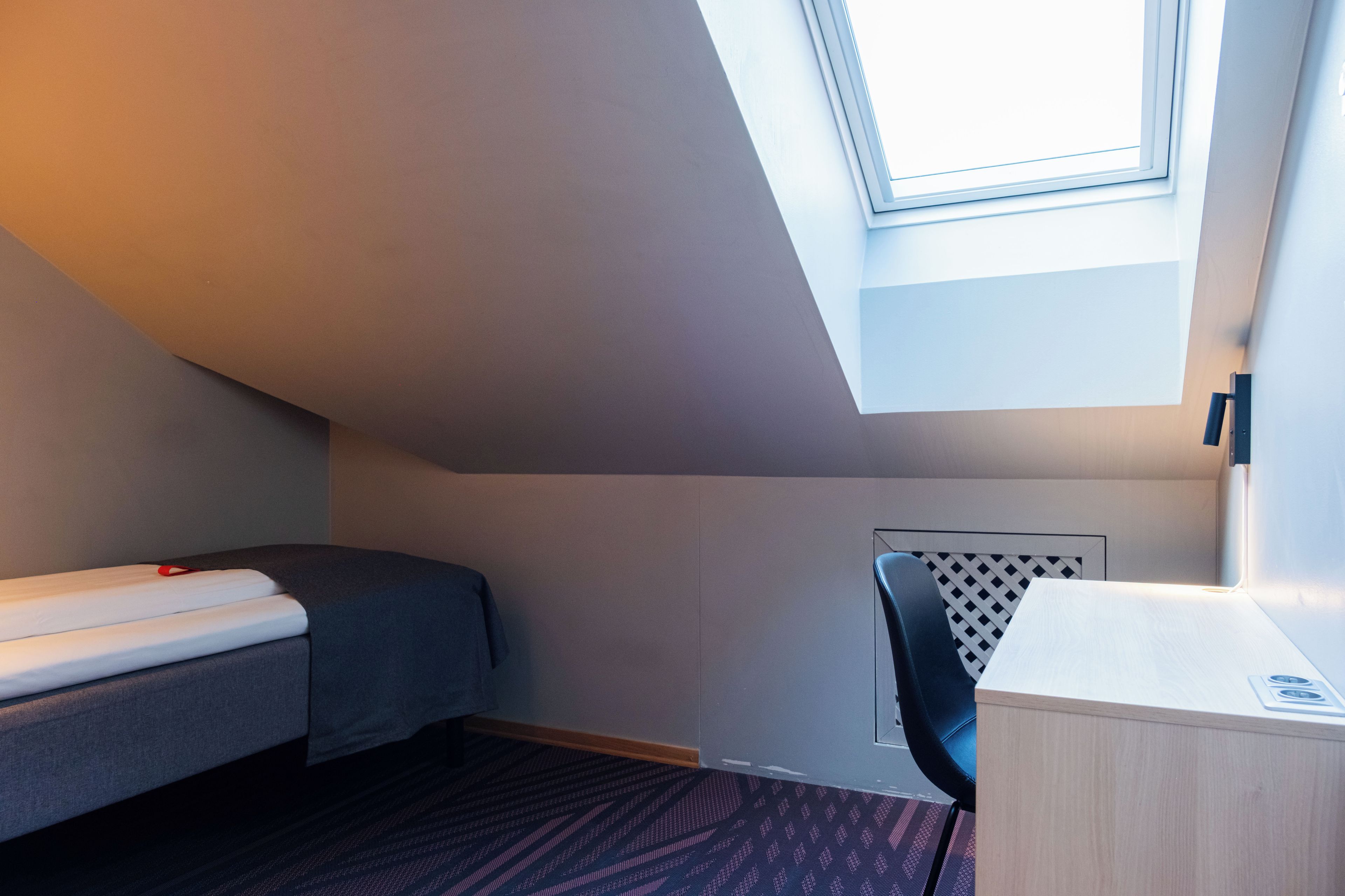 A small, compact room with a sloped ceiling, a single bed, a desk, and a chair.