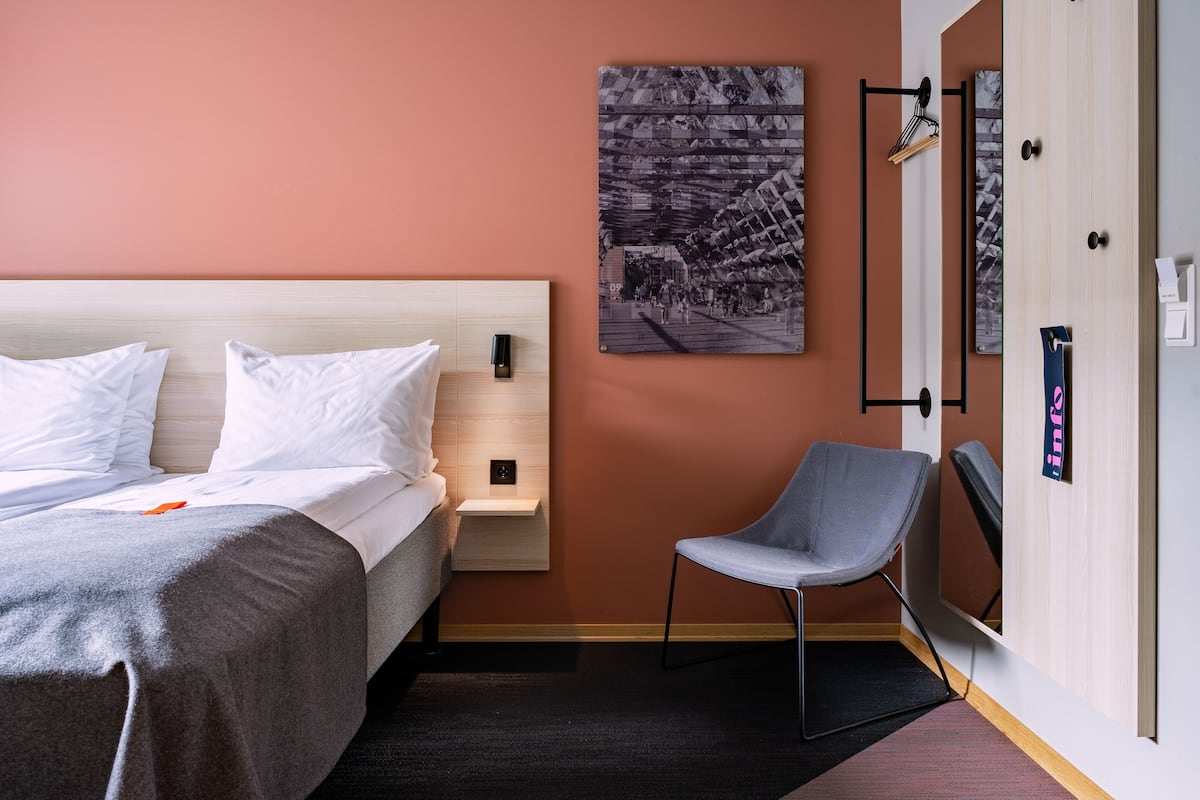 Hotellihuone, jossa on sänky, tuoli, seinälle ripustettu taideteos ja ovi, jossa on kyltti.