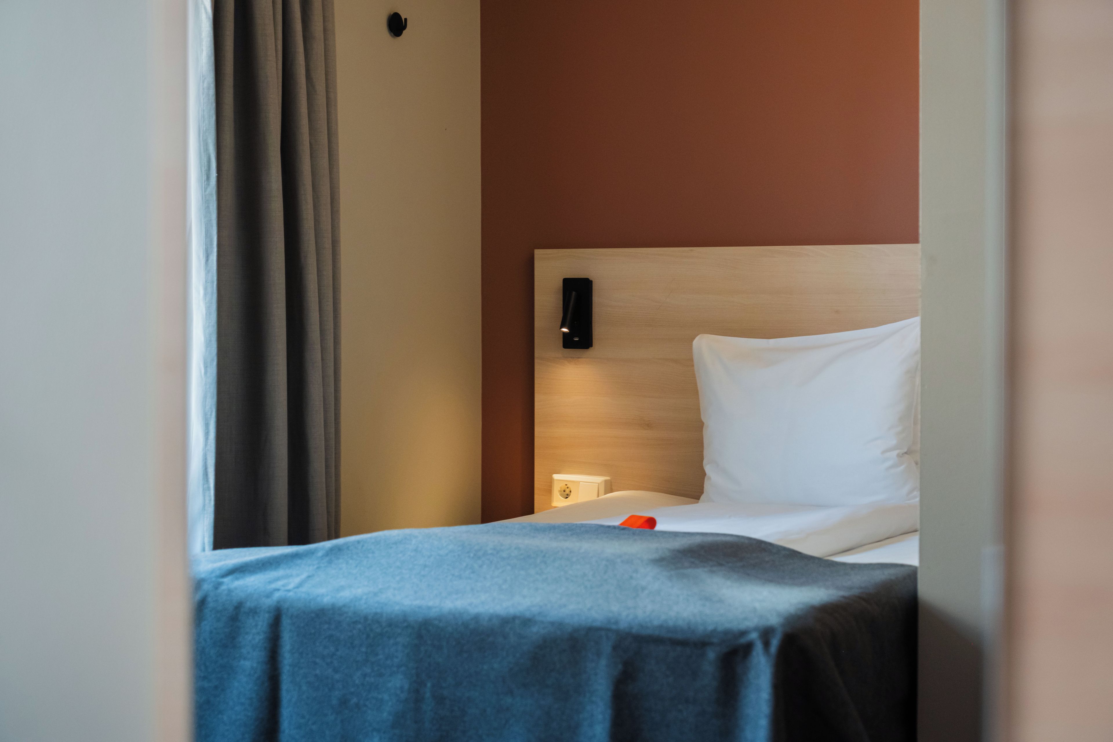 A hotel room with a bed, a bedside table, a wall-mounted phone, and a curtain.